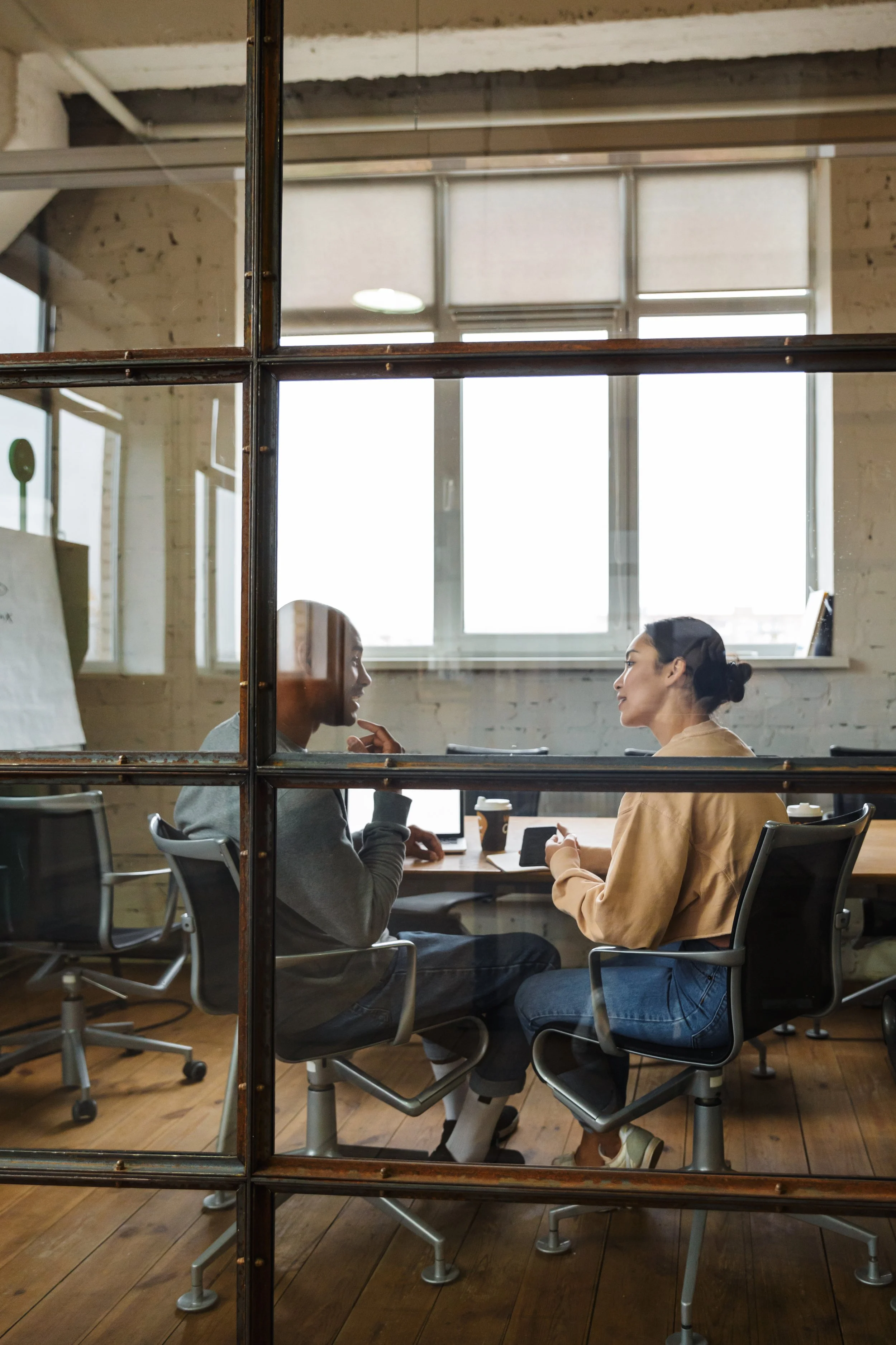 Two people having a conversation in an office, seen through a glass window with metal framing. The woman is wearing a beige jacket, and the man has a gray sweatshirt. They are sitting at a wooden table with coffee cups and a phone on it, in a bright room with large windows.