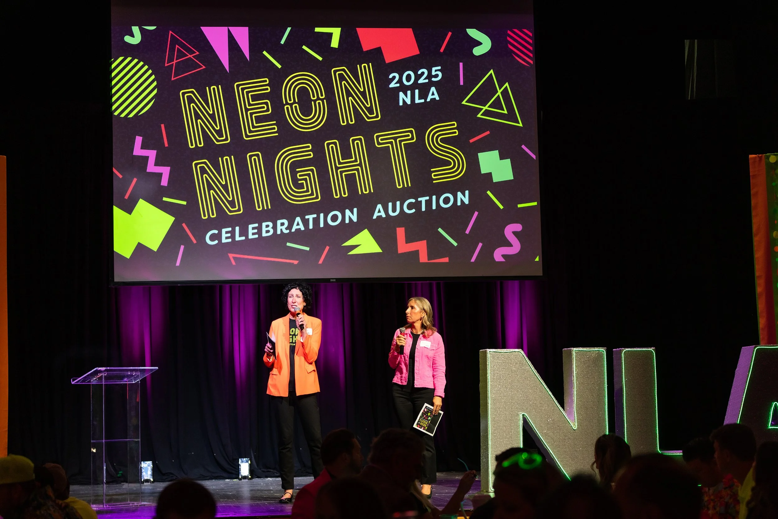 two ladies in neon blazers announcing on stage at auction event