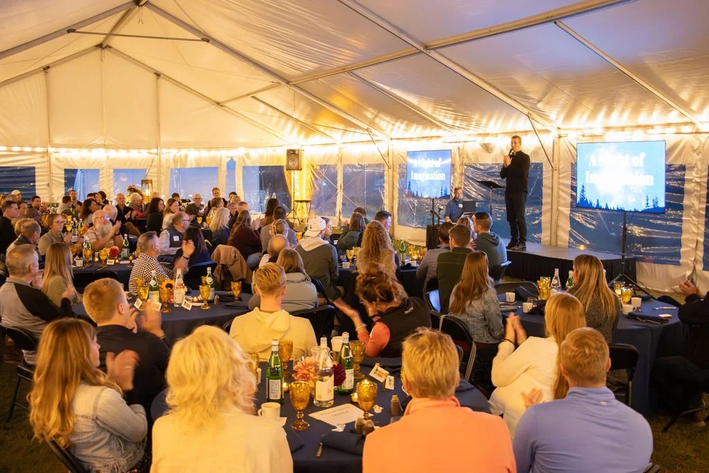Guests seated inside a tent with lights strung around the perimeter listening to a speaker on stage