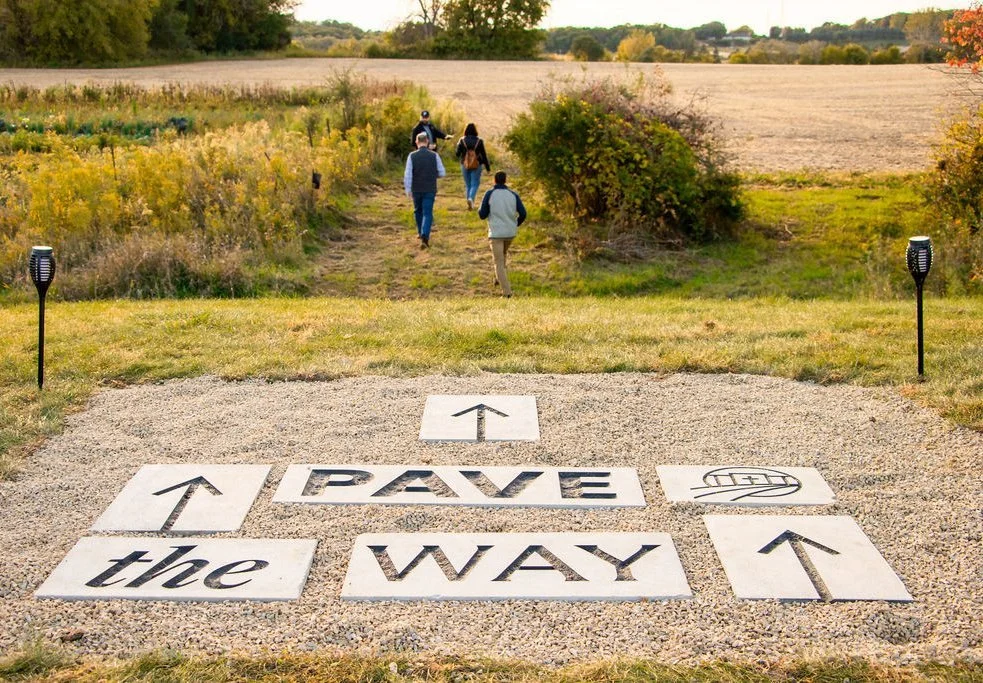 7 custom designed and created paver stones laid in the ground with arrows and the words Pave the Way leading out to field with people walking