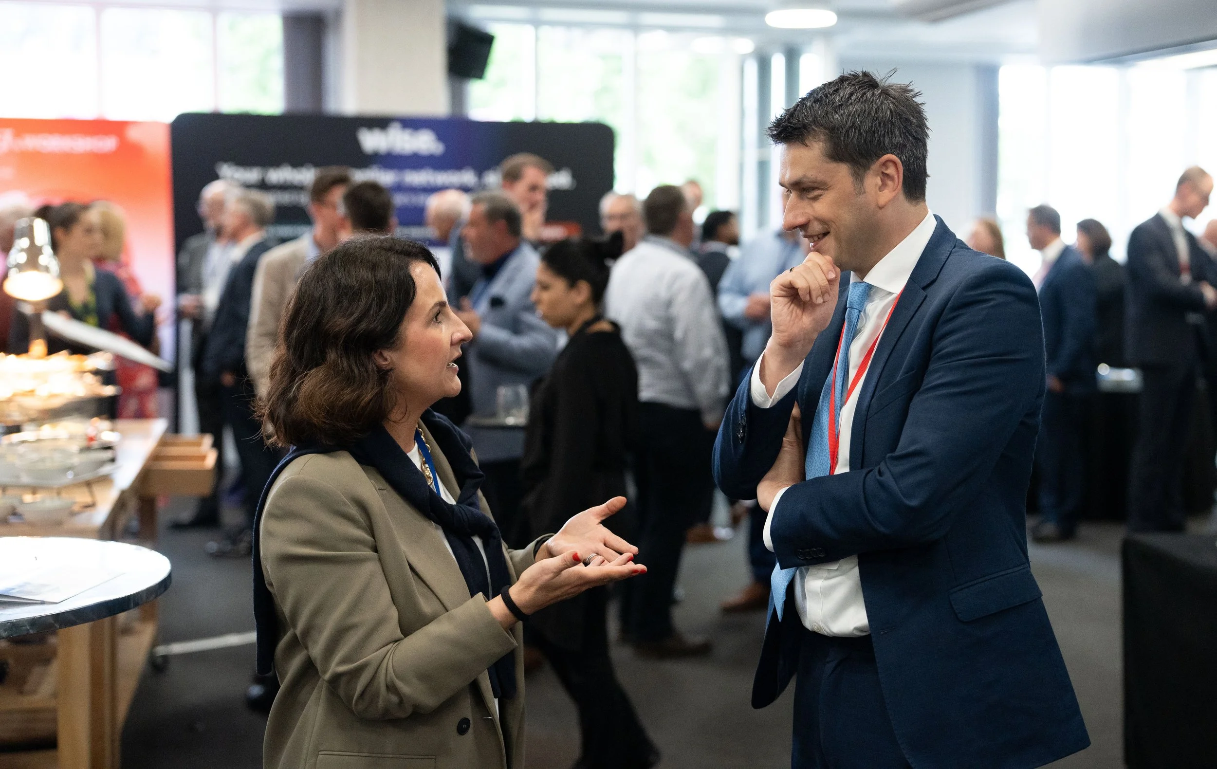 Two people, a woman and a man, are engaged in conversation at a professional conference, with other attendees in the background.