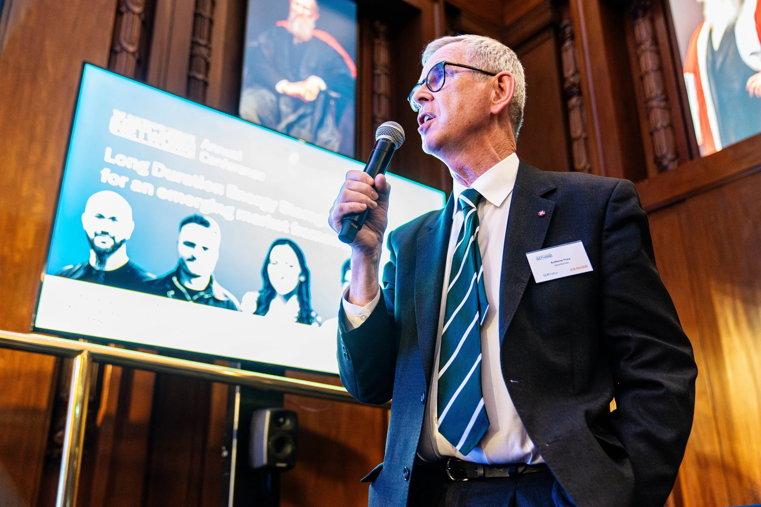 Older man with glasses, dressed in a dark suit and striped tie, speaking into a microphone during a presentation at a conference. Behind him is a large screen displaying a slide with portraits of three people and a light blue background.
