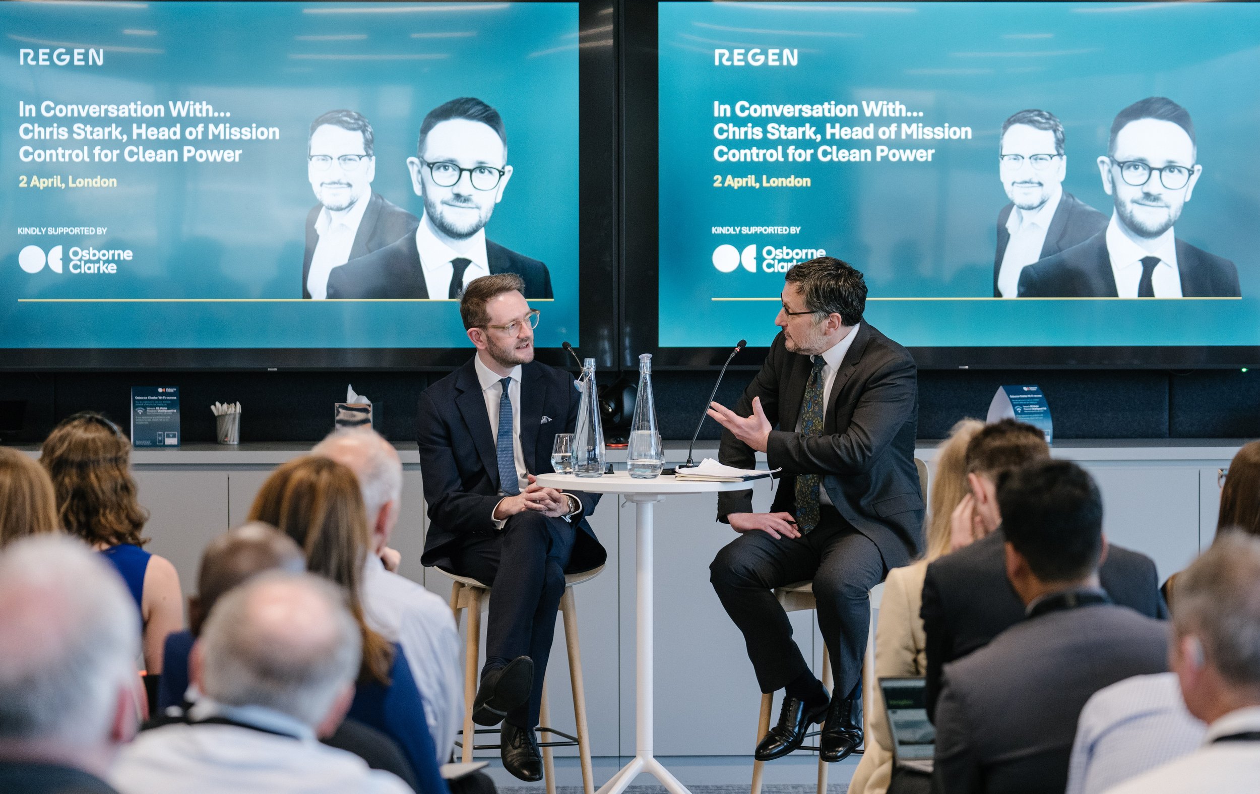 A man in a dark suit and glasses is speaking into a microphone during a panel discussion, while another man in a suit listens attentively. Both are seated at a round table with water bottles. An audience of diverse people watches the discussion, with two large screens behind them displaying a promotional graphic for an event titled 'In Conversation With Chris Stark, Head of Mission Control for Clean Power,' held on April 2 in London, supported by Osborne Clarke.