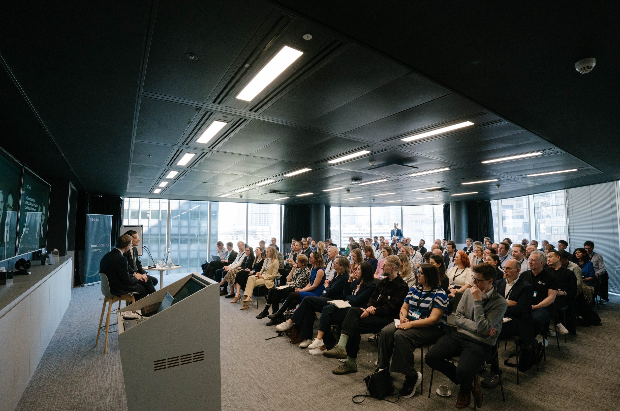 A large group of people attending a conference or seminar in a modern, high-rise office space with large windows.