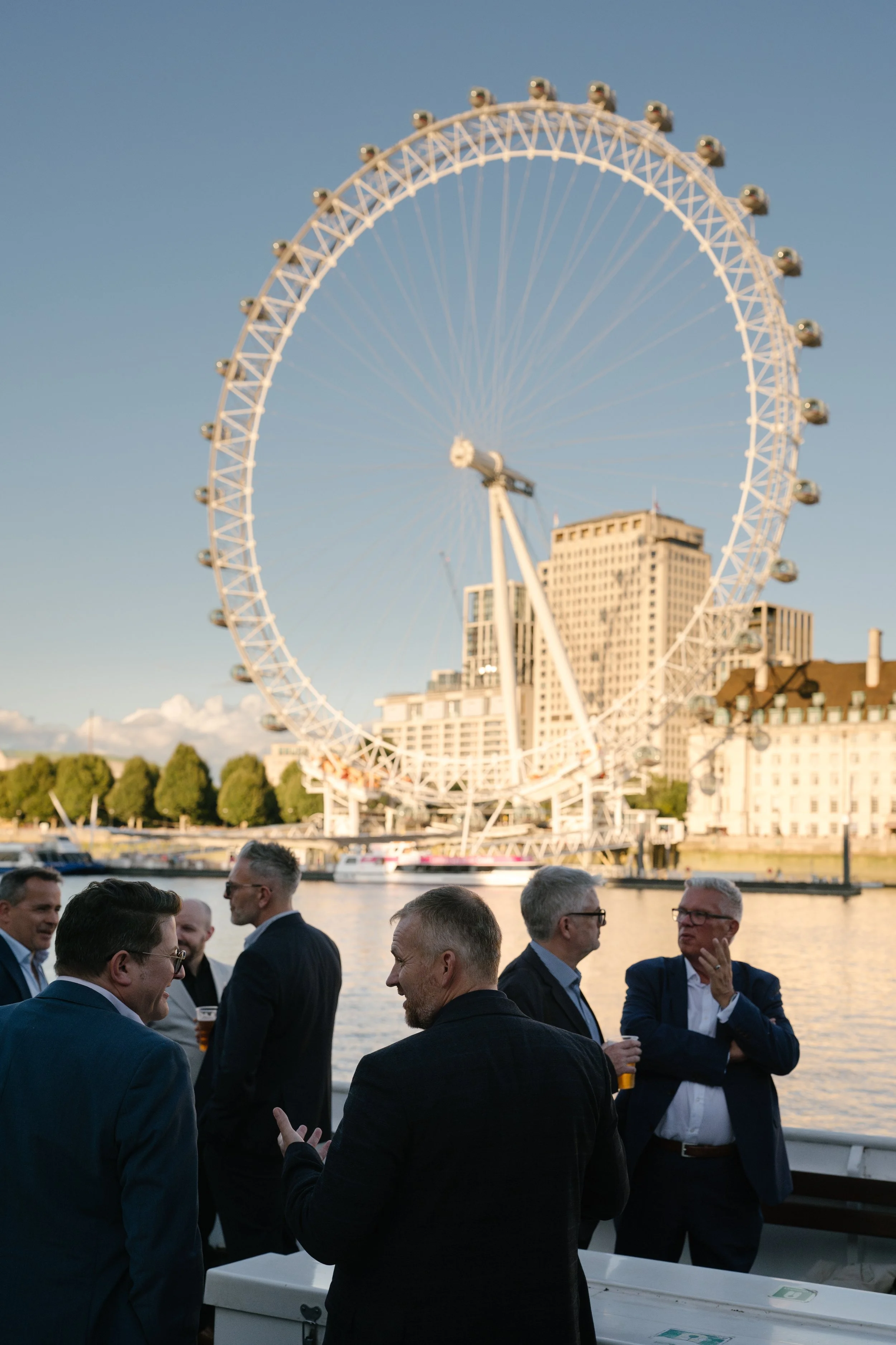 A group of men in suits, attending a business event, are talking in front of the London Eye