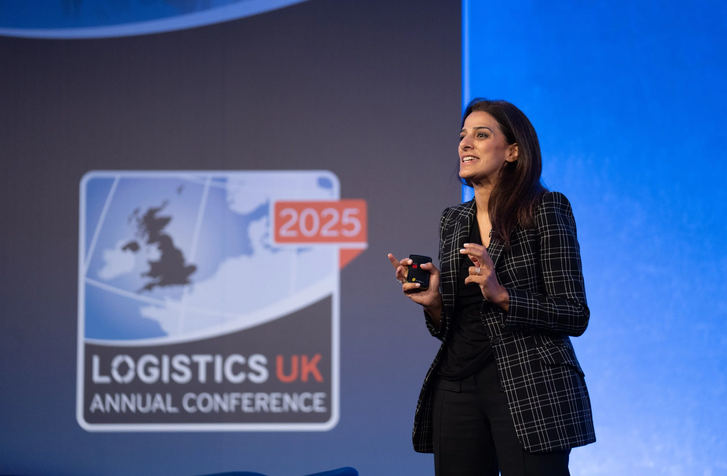 A woman speaking at a corporate conference with a sign that reads 'LOGISTICS UK ANNUAL CONFERENCE 2025' behind her.