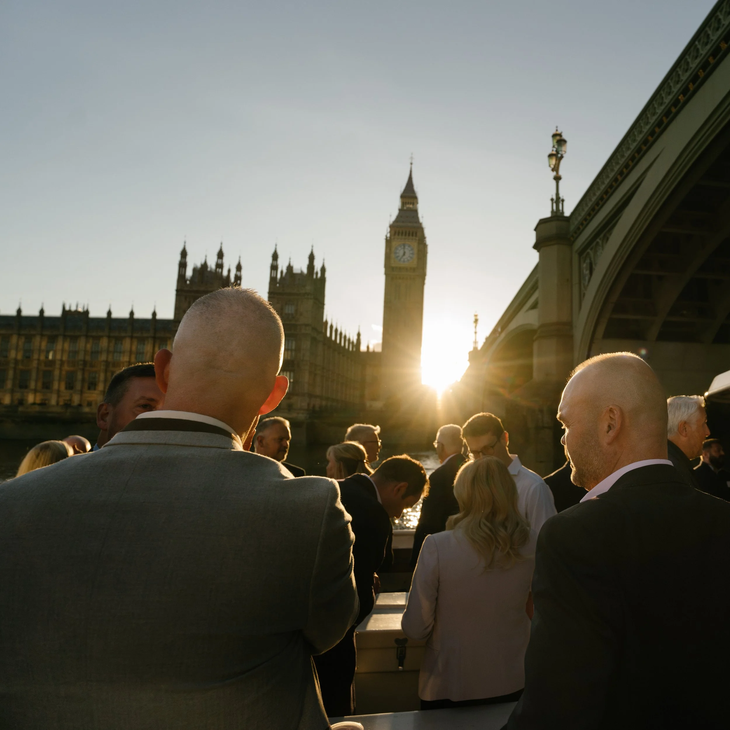 People gathered outdoors at a corporate event near Westminster Bridge with Big Ben in the background at sunset.