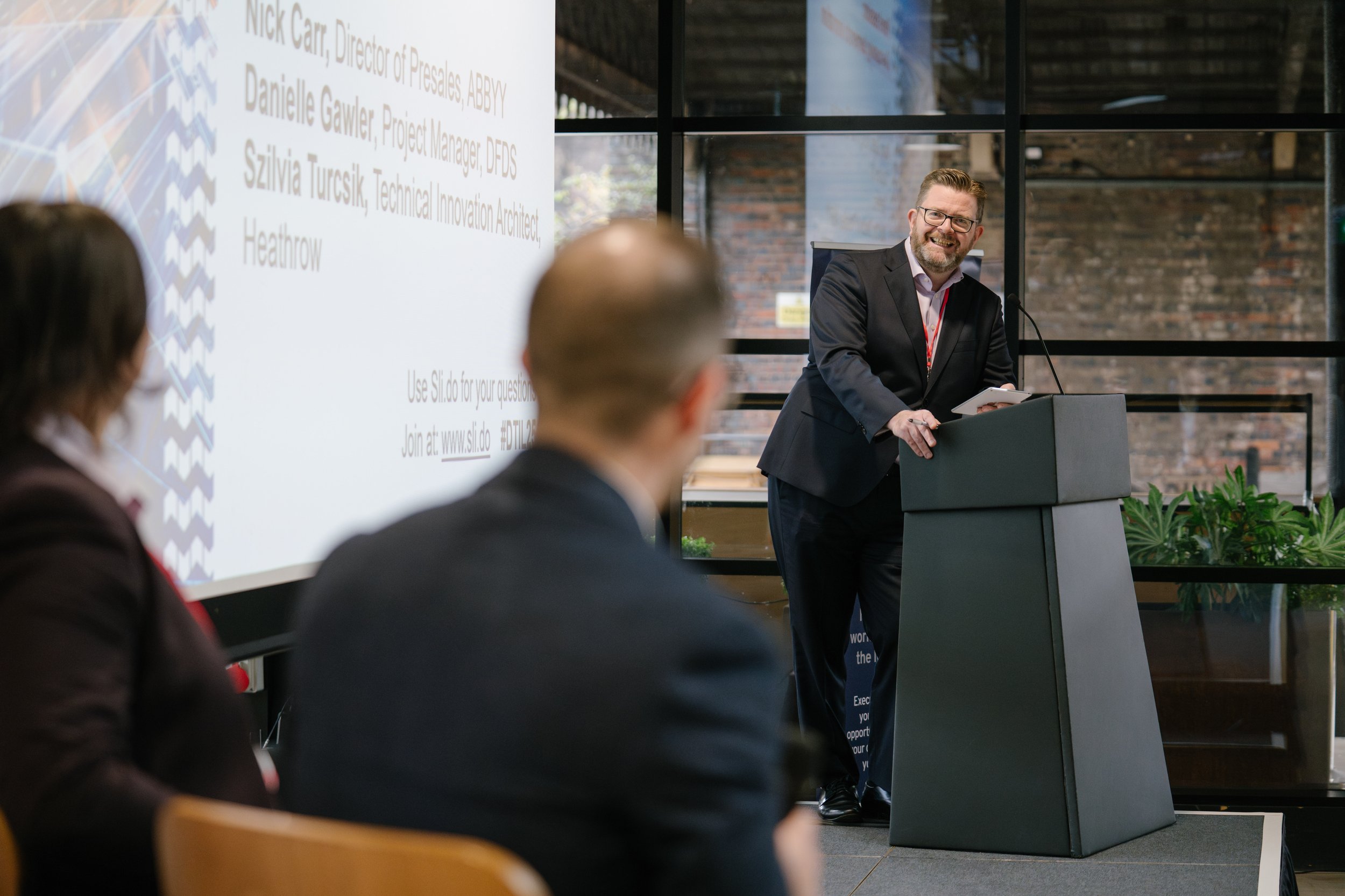 A man with a beard is speaking at a corporate event and is smiling at a member of the audience.