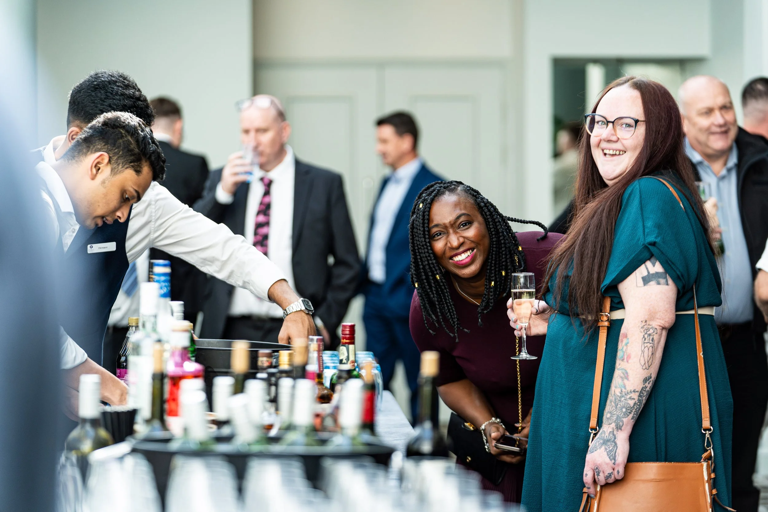 People socialising at an event, with two women smiling and holding a glass of champagne in focus, in a room with other attendees in the background.