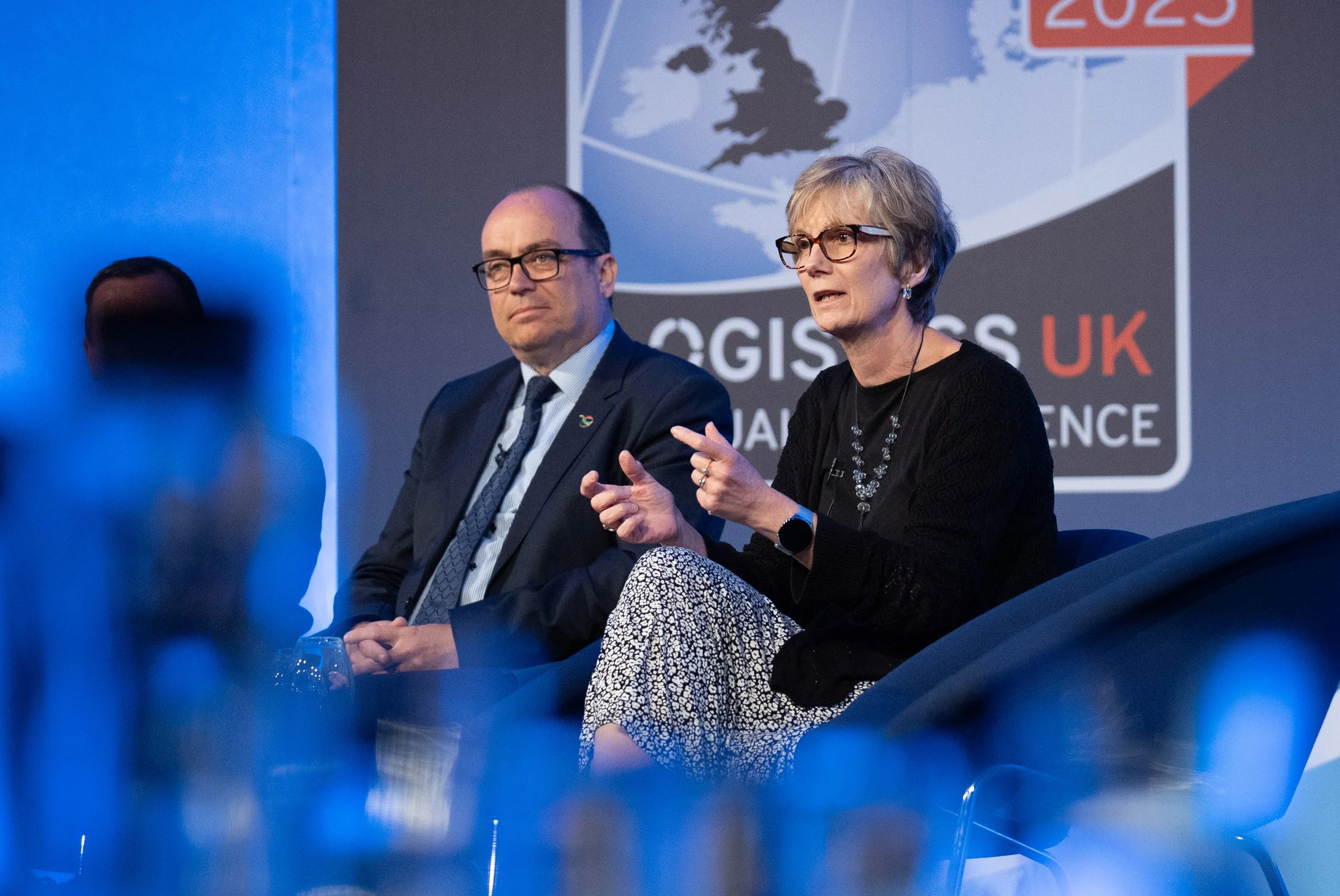 A woman and a man seated on a stage during a conference. The woman is speaking and gesturing with her hands, wearing glasses, a black top, and patterned pants. The man, dressed in a suit and tie, is listening with a neutral expression. There is a blurred figure in the foreground, and a large screen behind them displaying a logo for a UK logistics conference with a map of the United Kingdom.