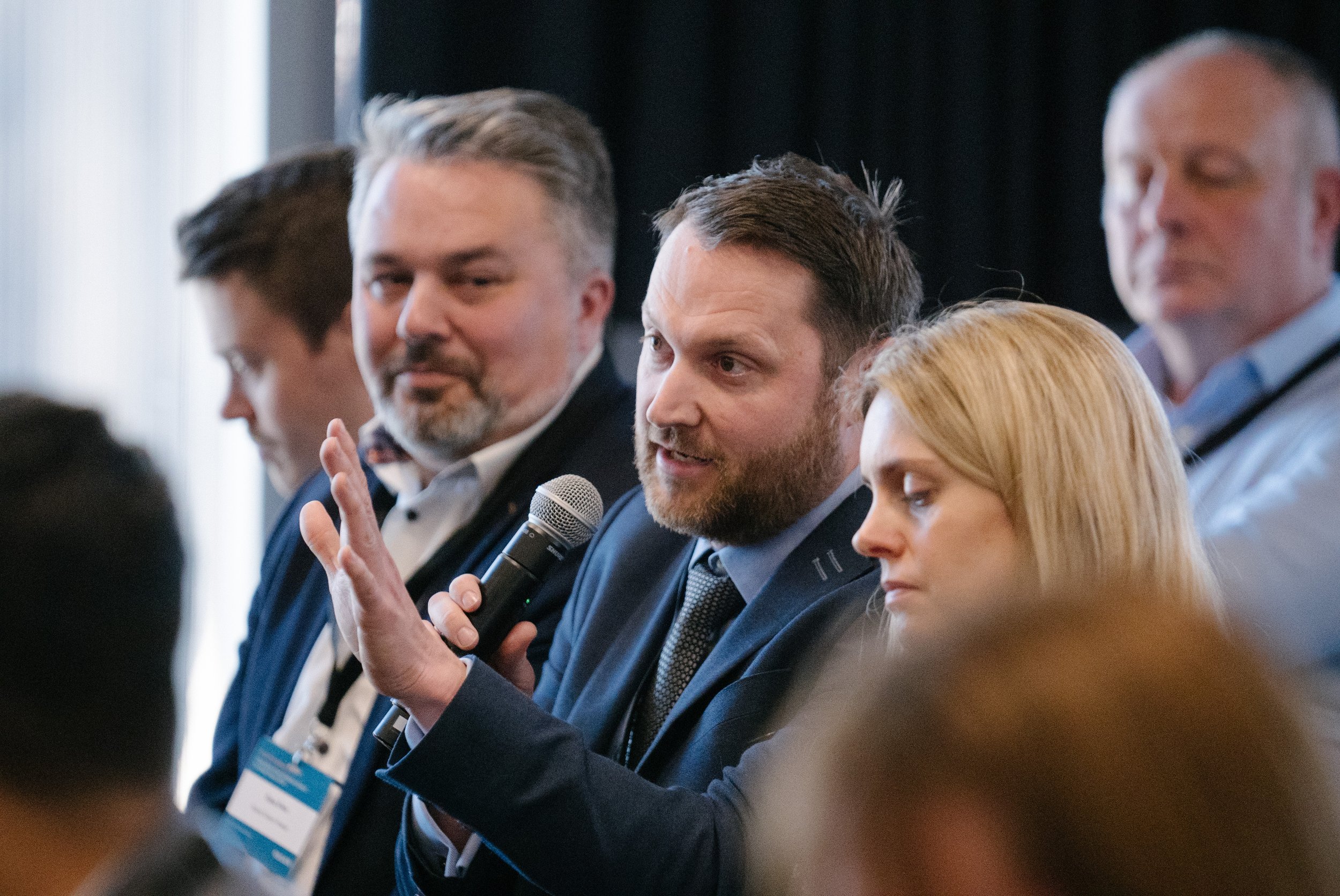 A group of professionally dressed people attending a business conference, with one man holding a microphone and speaking.