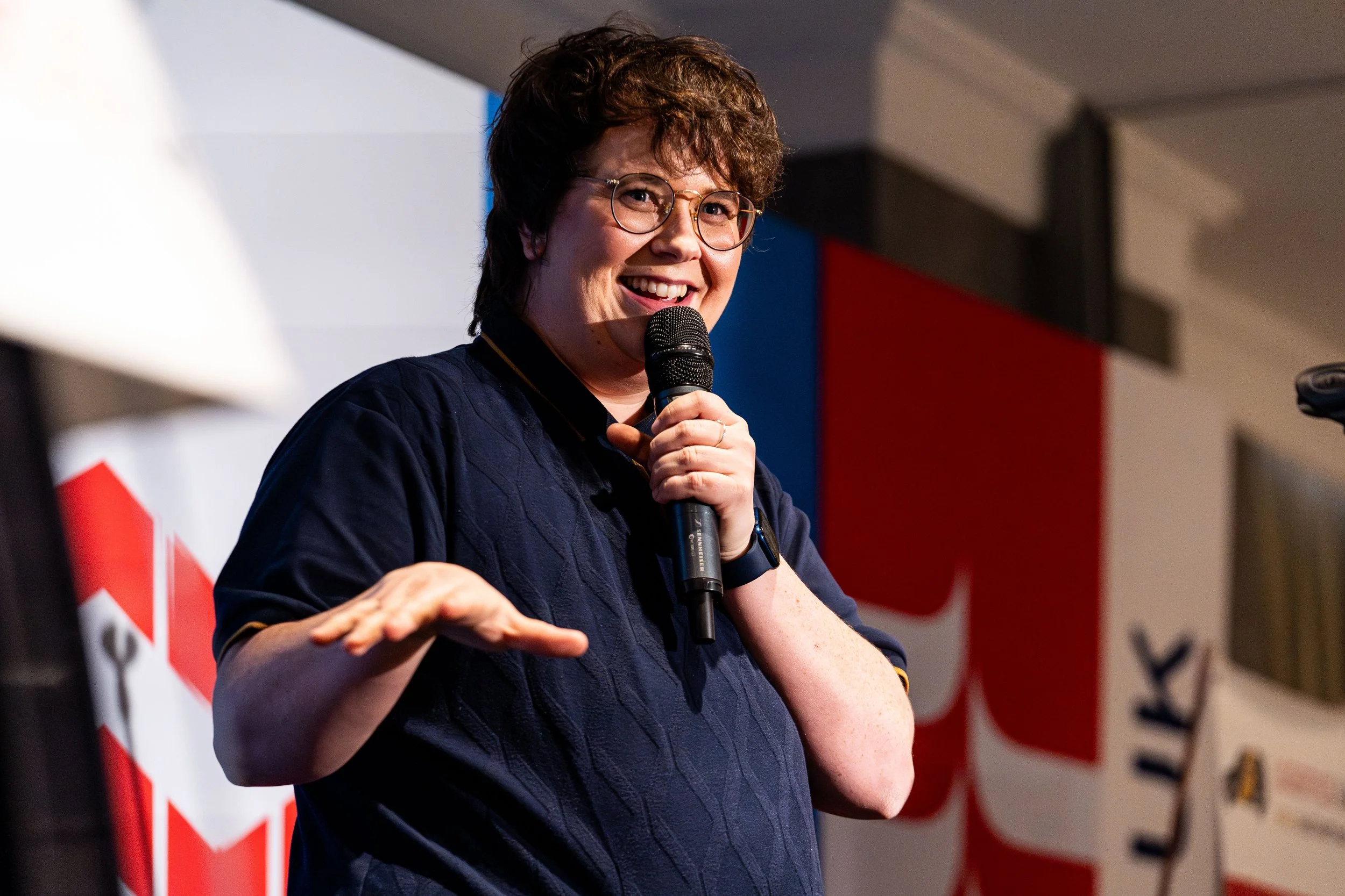 A woman with short curly hair and glasses smiling while holding a microphone during a speech. She is wearing a navy blue shirt.