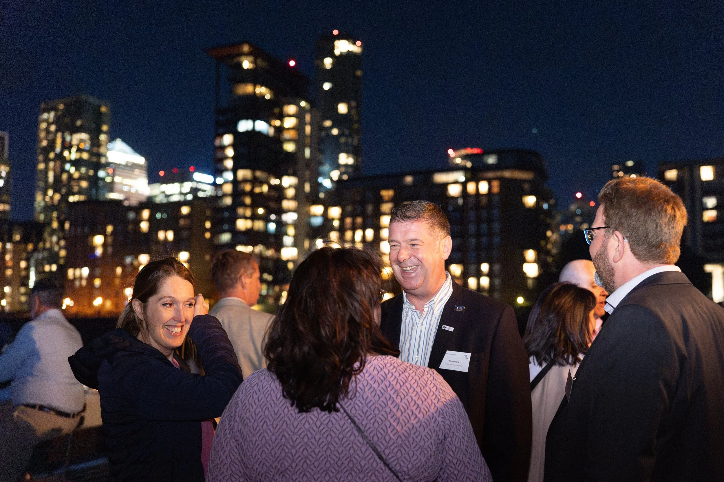 People at a nighttime corporate event on a rooftop with city skyline in background, engaging in conversations and smiling.