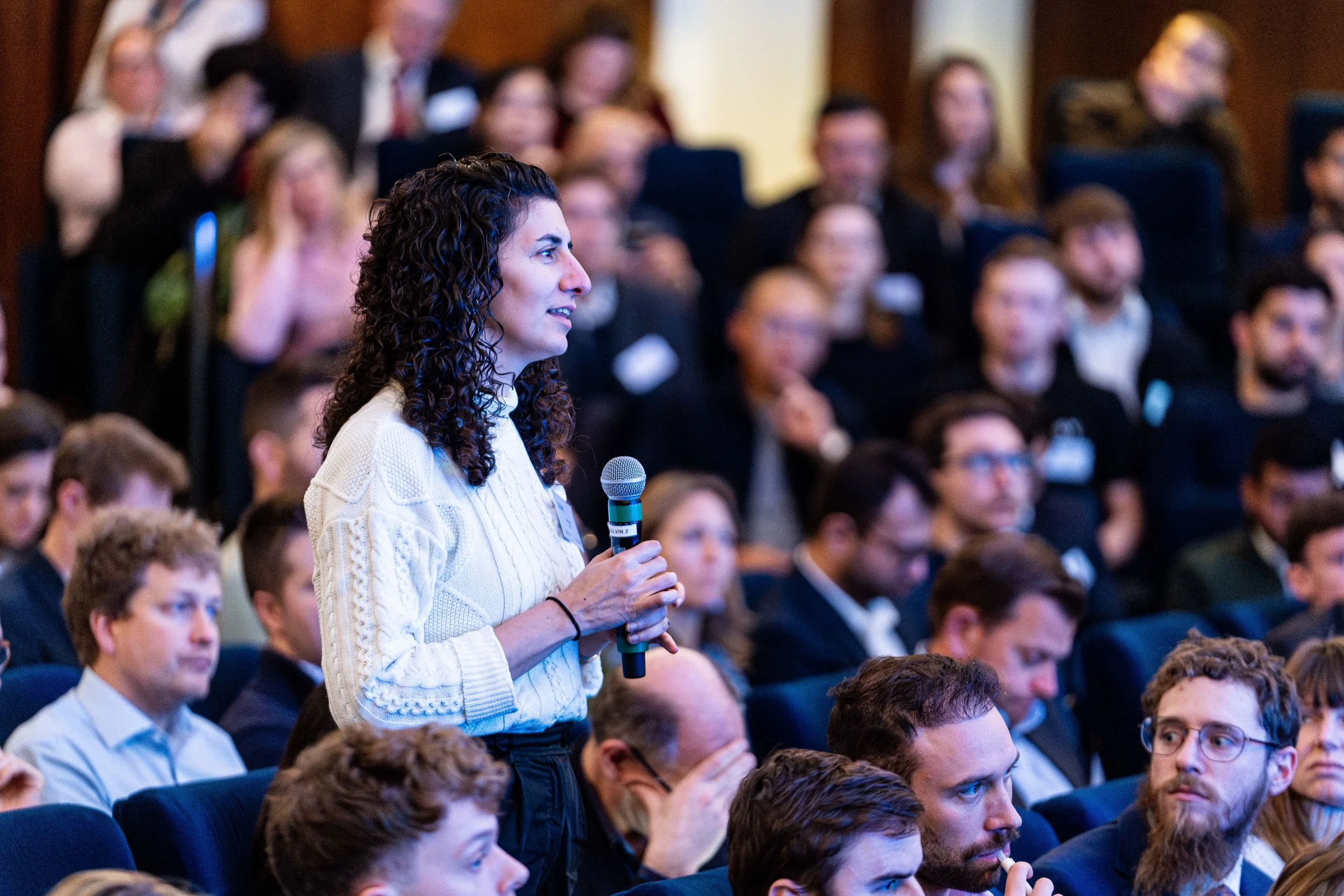 A woman with dark, curly hair in a white sweater holds a microphone while attending a business conference with a seated, attentive audience in a formal setting.