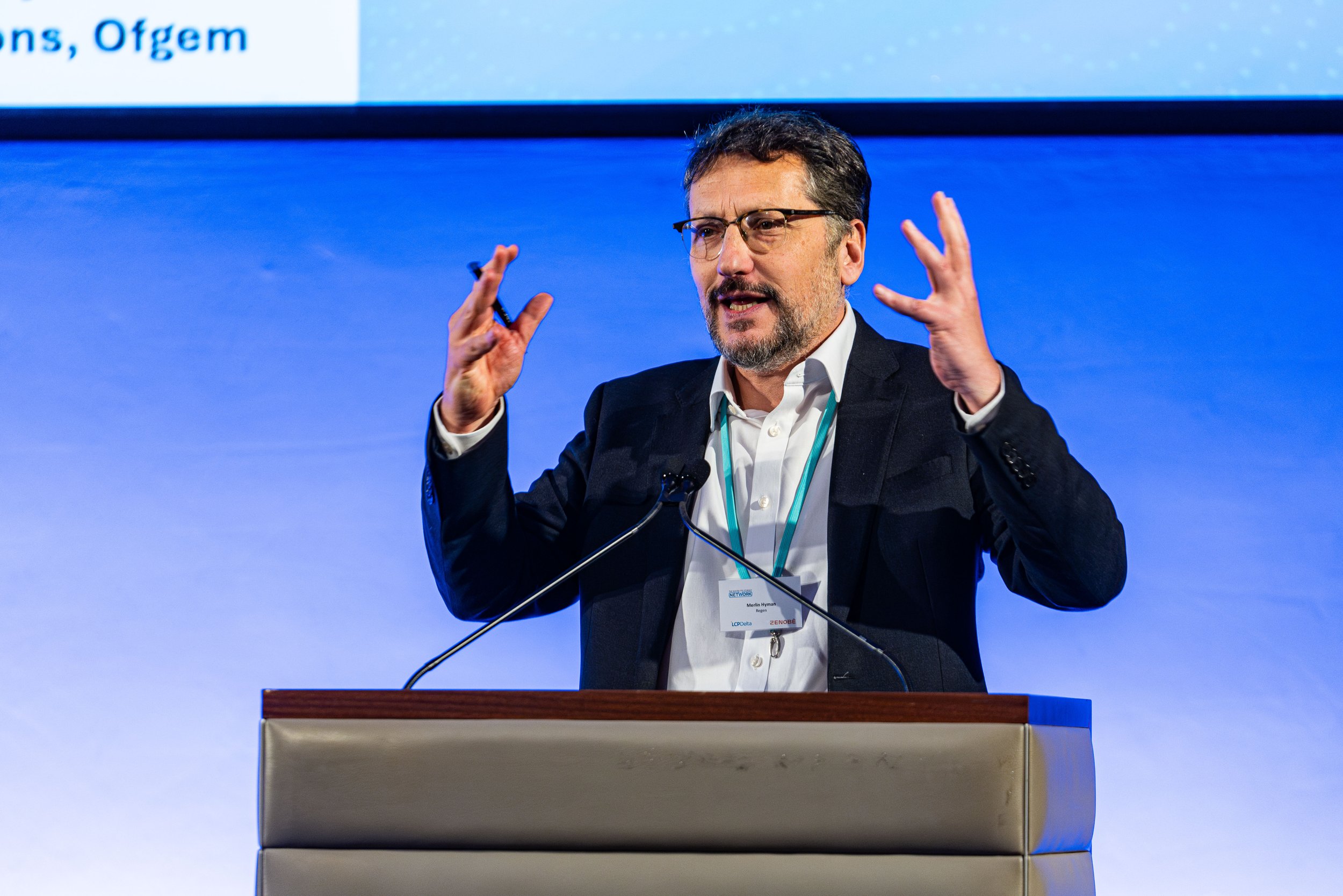A man wearing glasses and a dark suit jacket giving a presentation at a conference, standing behind a podium with a microphone, gesturing with his hands.