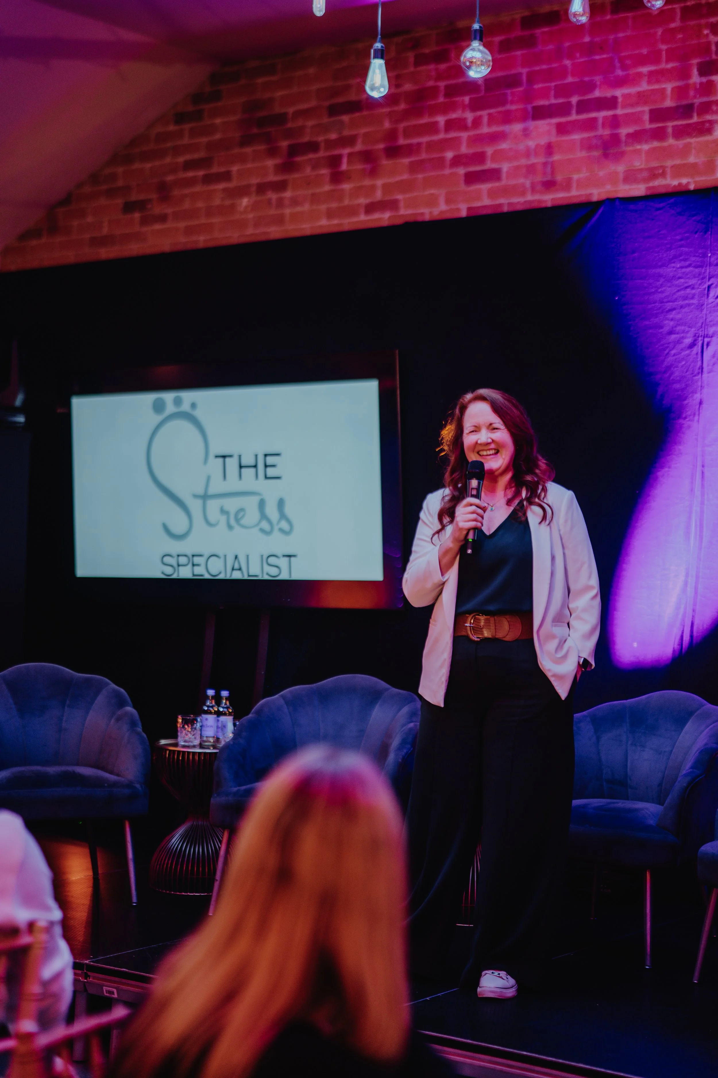 A woman with curly red hair, laughing, standing on a stage with a microphone - backlit by a screen with The Stress Specialist logo on it