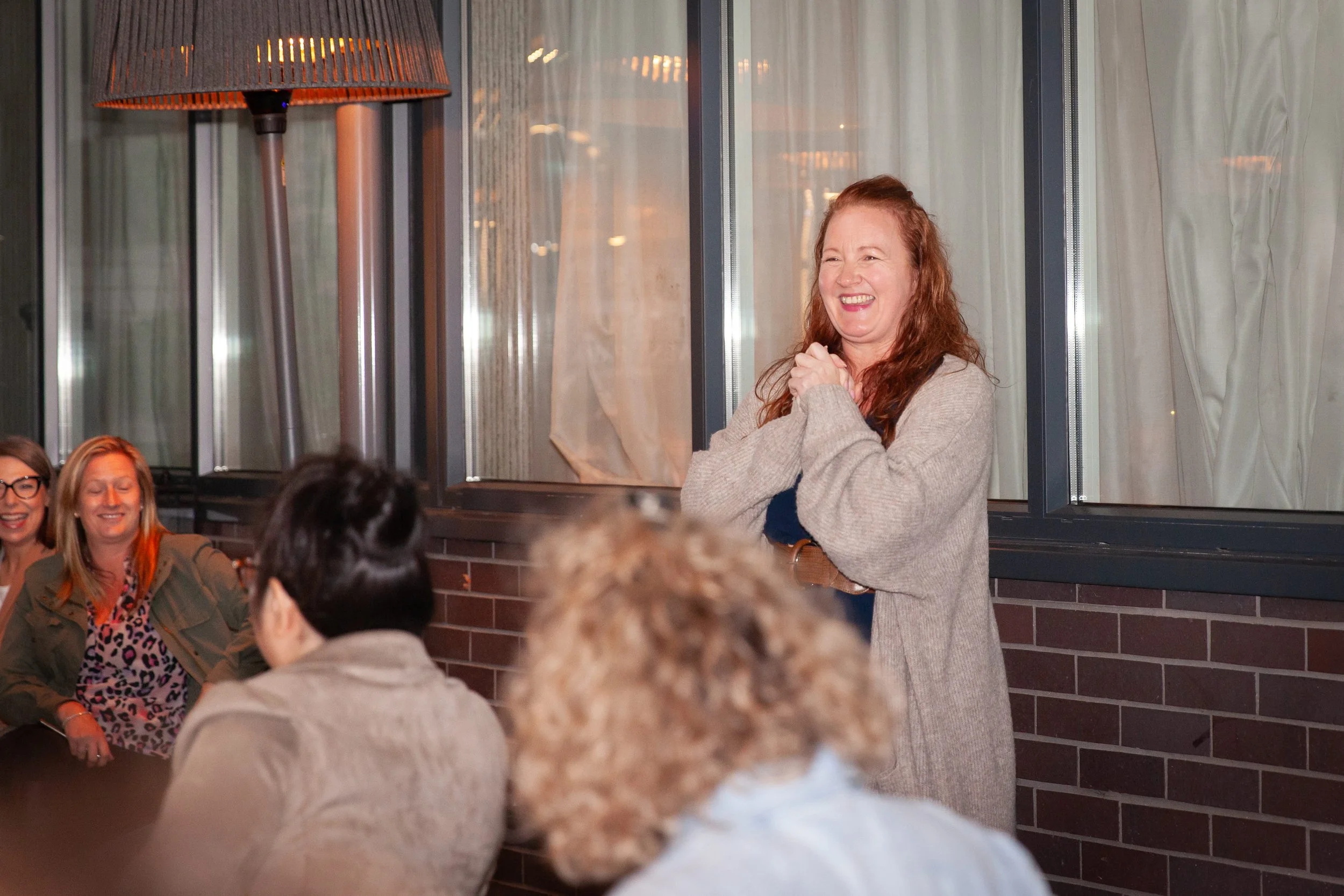 A woman with red hair and a beige cardigan smiling and standing with her hands clasped in front of her chest, speaking to a group of women seated around a table in a room with large windows and brick wall.
