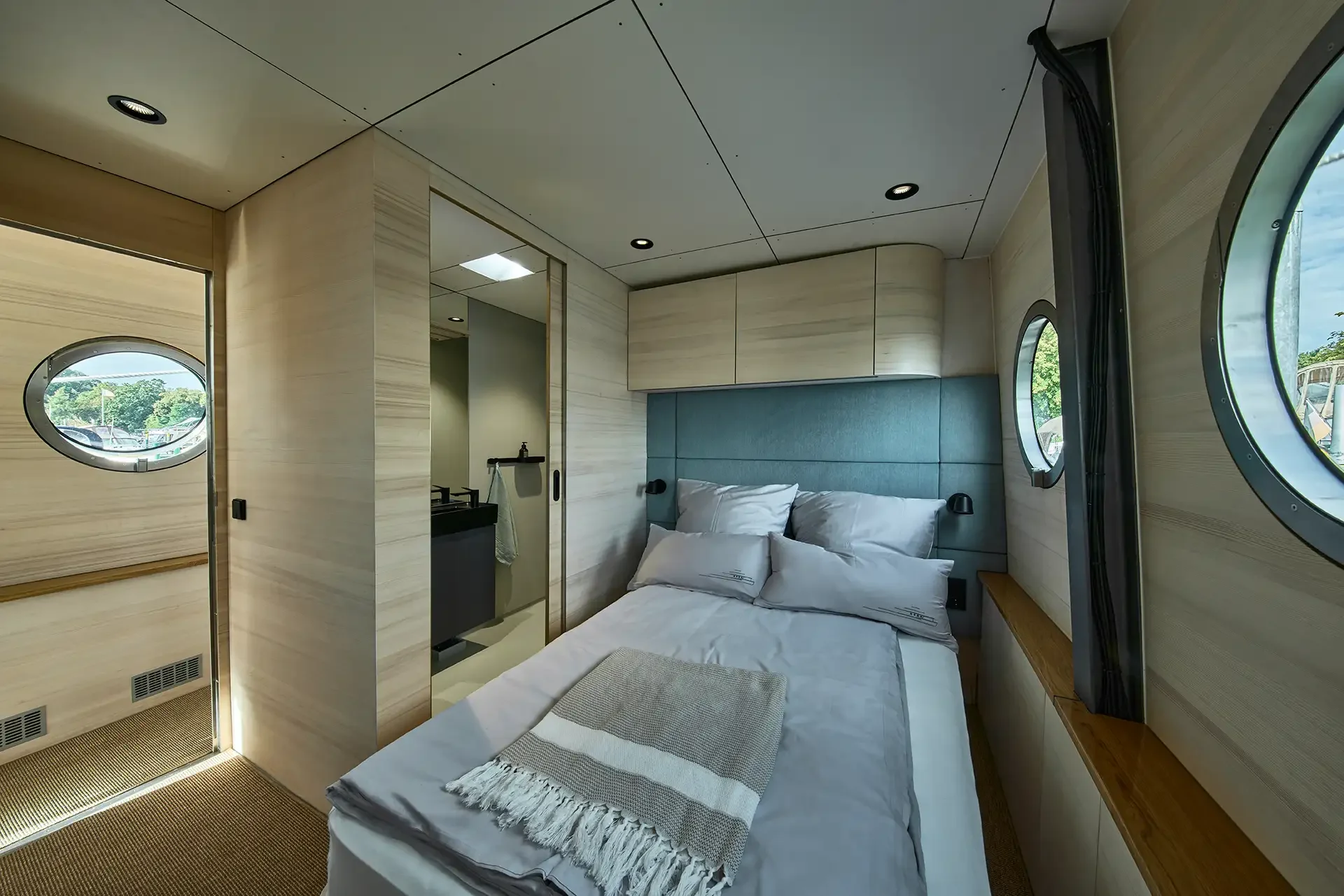 Kleines Schlafzimmer in einem modernen Hausboot mit Doppelbett, blauer Kopfteil, vier Kissen, einer Decke, seitlichen runden Fenstern, Holzverkleidungen und einem kleinen Waschbeckenbereich.