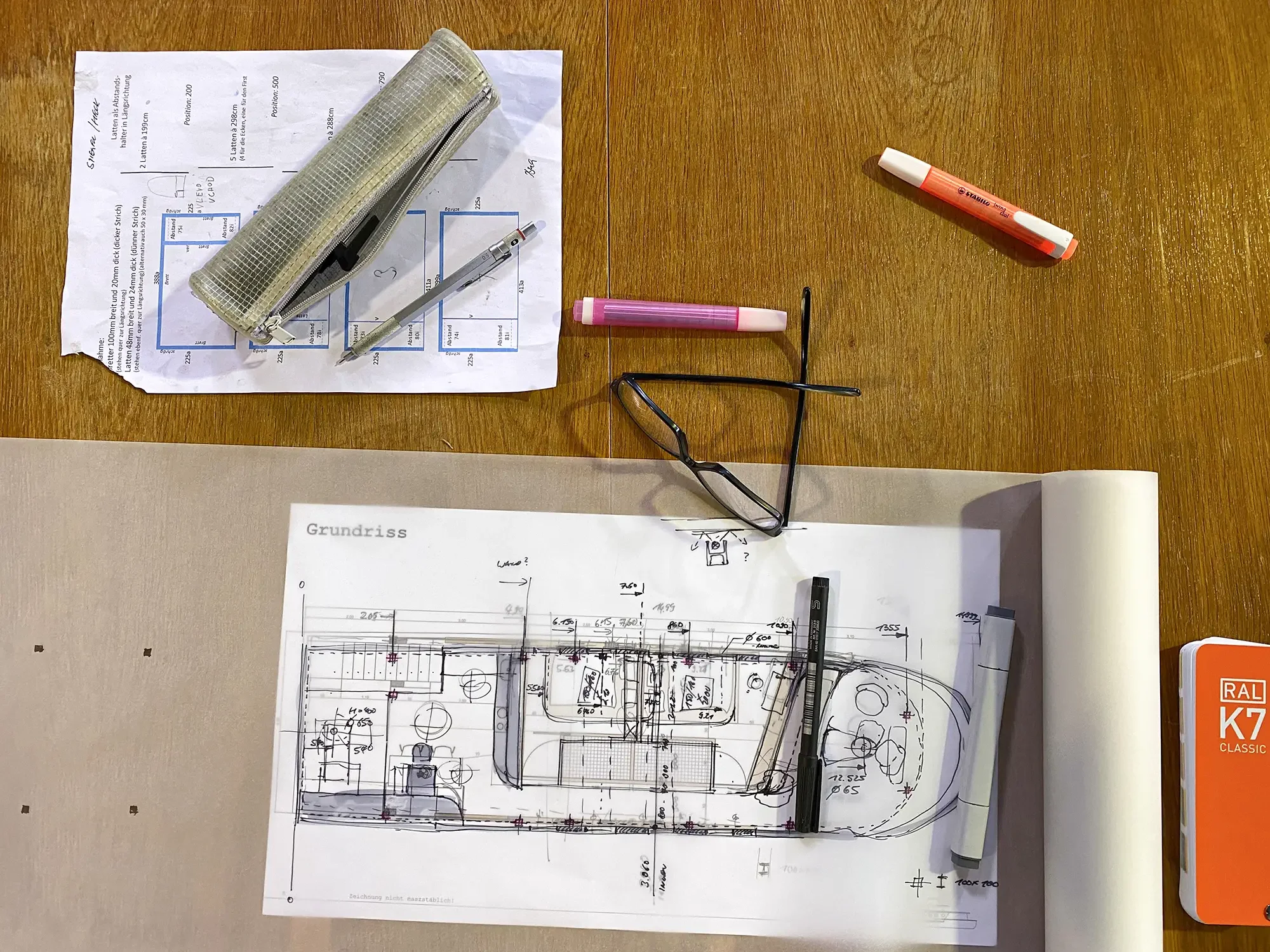 Architekturskizze auf einem Tisch mit Bleistift, Marker, Brille, Pinnwand-Klammer, getrocknetem Marker, Schreibwaren und Papierrollen.