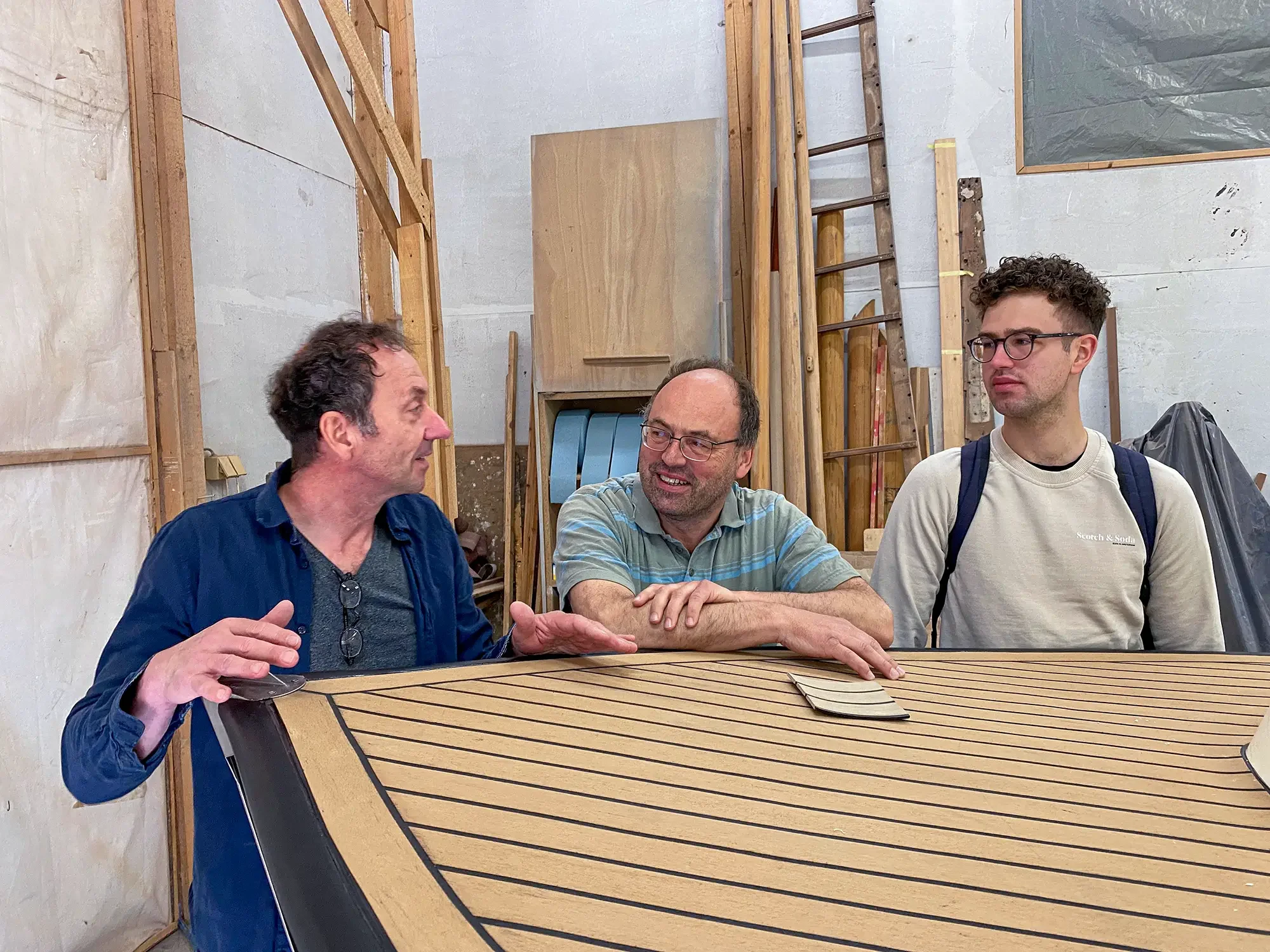 Drei Männer in einer Werkstatt, die um ein großes Holzstück mit schwarzen Linien sitzen und reden.