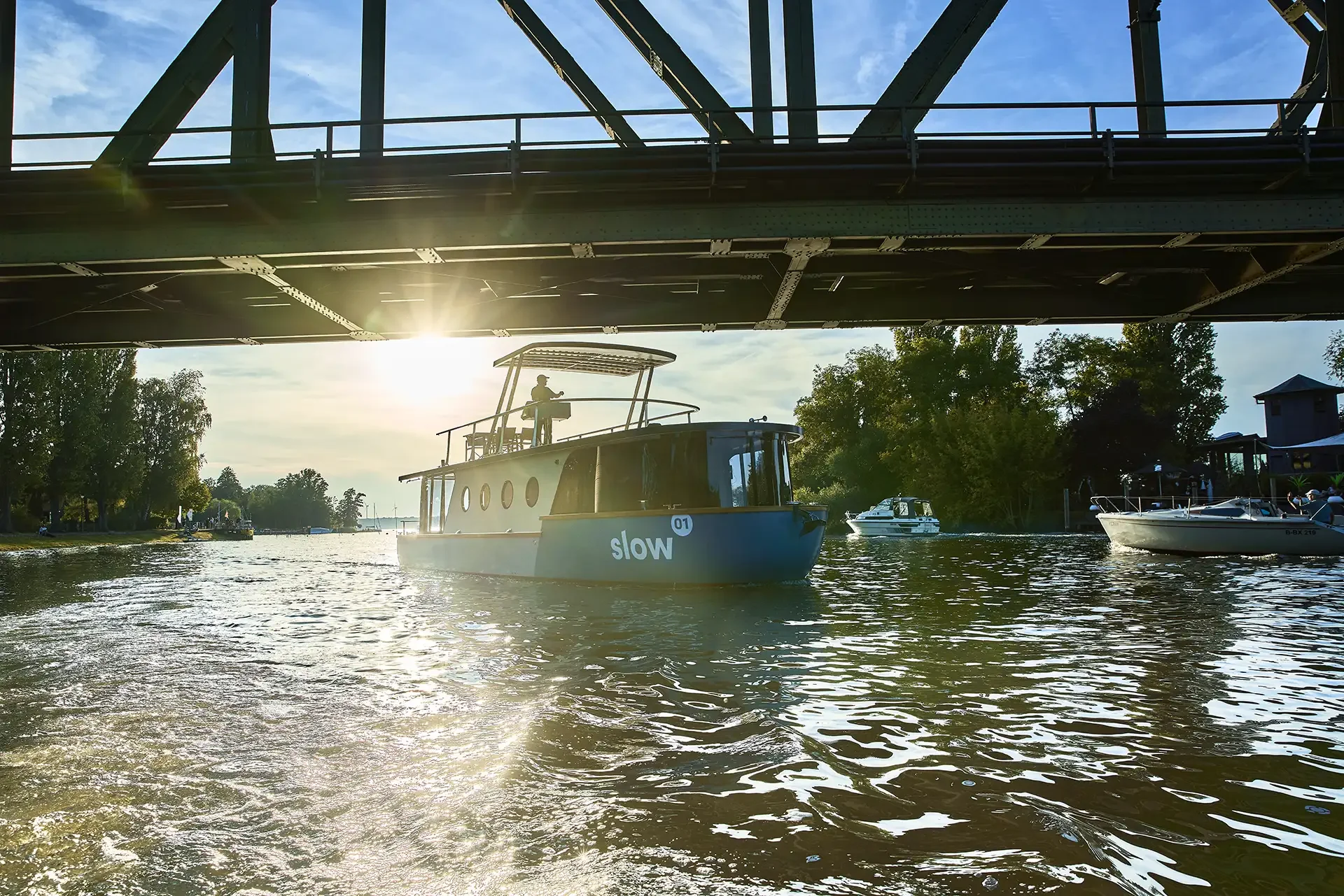 Boot auf einem Fluss, sichtbar unter einer Brücke, mit Sonnenlicht im Hintergrund, umgeben von Bäumen und weiteren Booten.