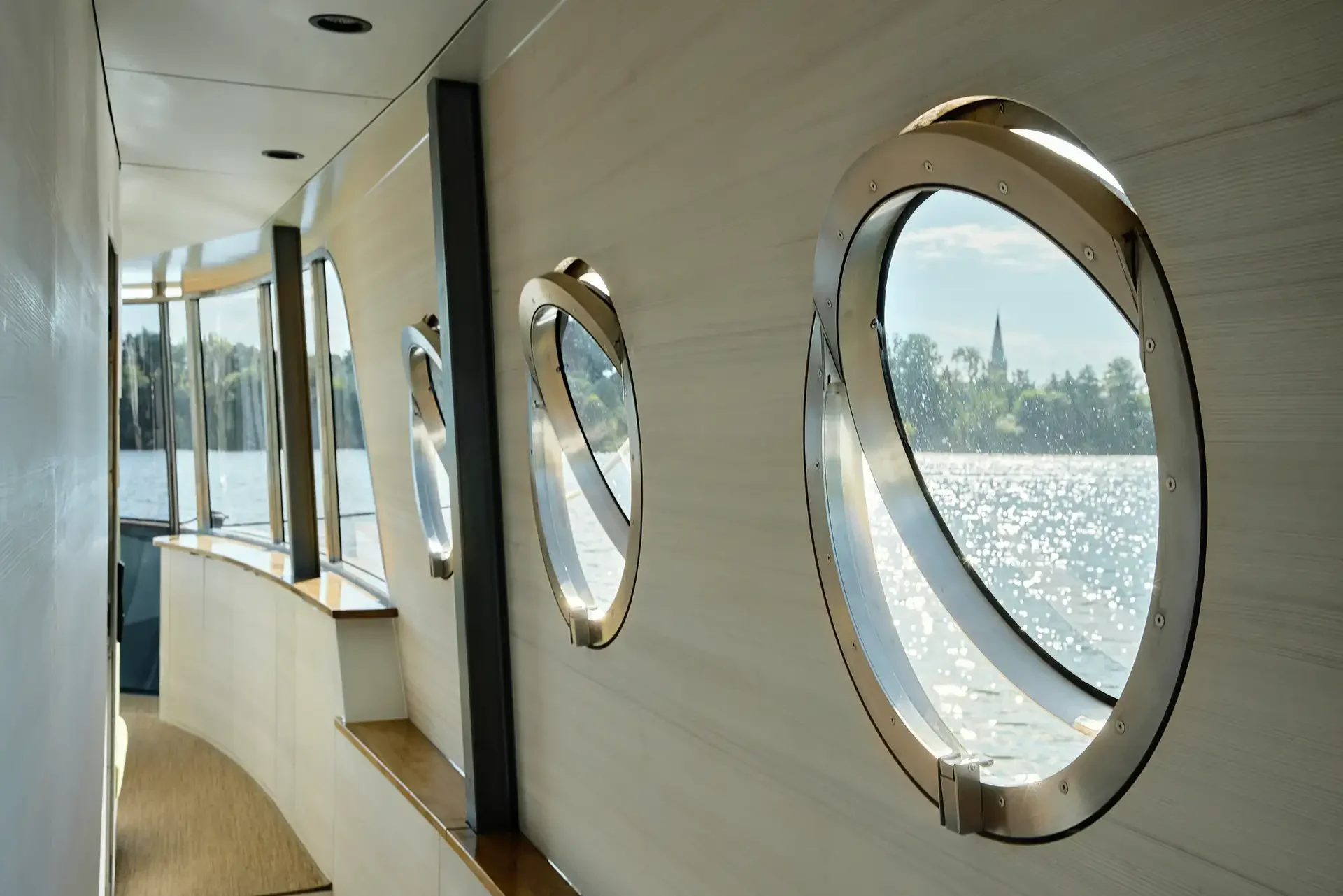 Innenansicht eines Schiffs mit runden Bootsfenstern, Blick auf einen Fluss oder See und eine grüne Landschaft mit Bäumen und einer Kirchturm im Hintergrund.