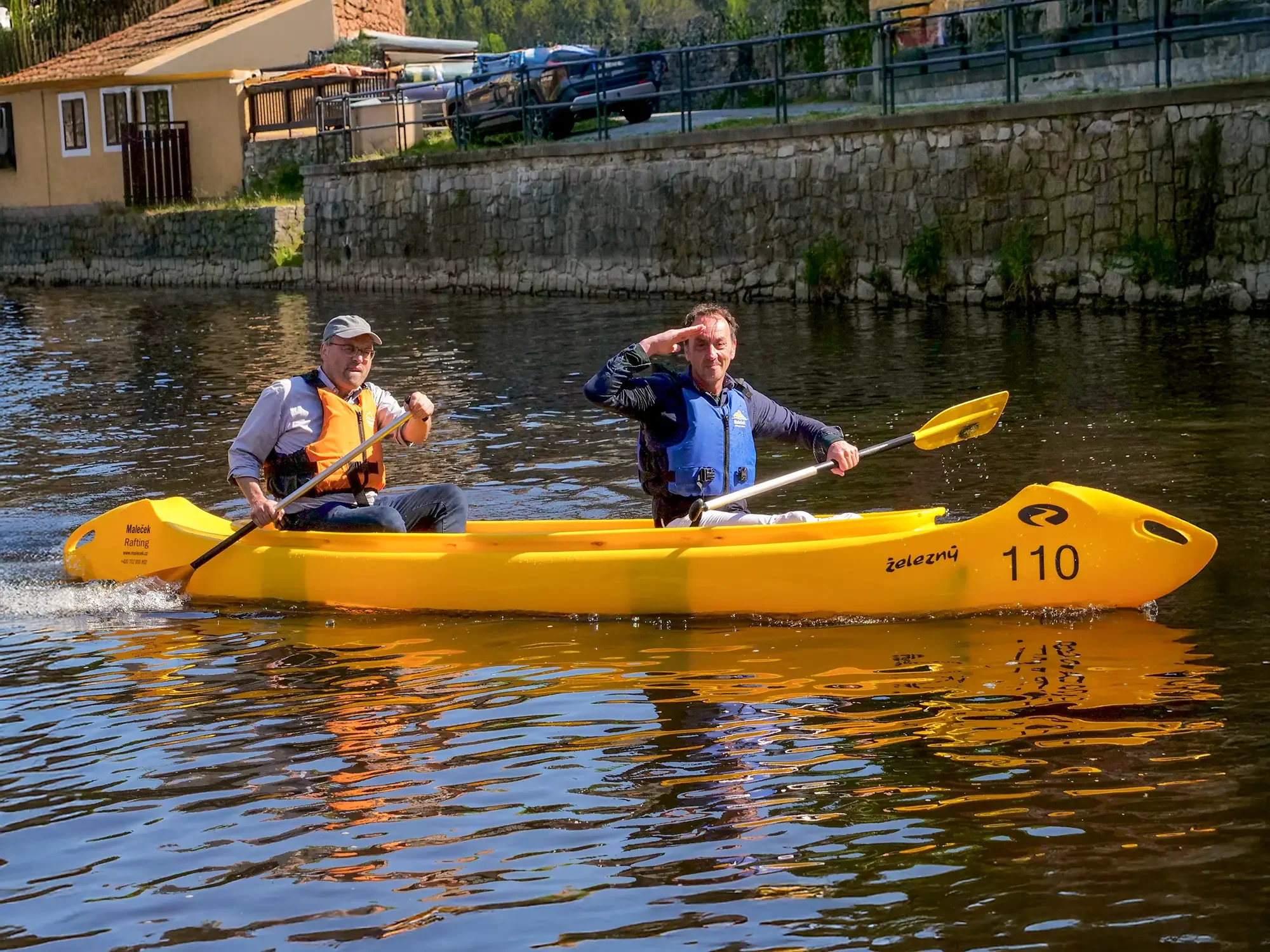 Zwei Männer paddeln in einem gelben Kajak auf einem Fluss. Einer trägt eine orange Schwimmweste und eine Mütze, der andere ein blaue Schwimmweste, während er salutiert.