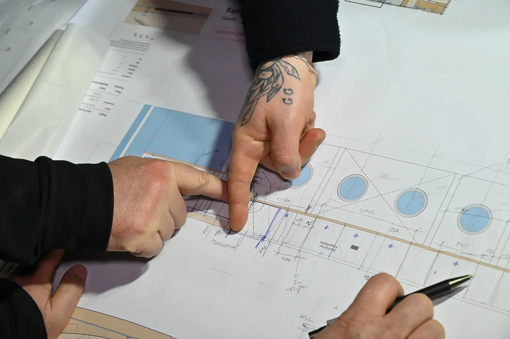 Zwei Menschen betrachten einen Bauplan. Eine Person zeigt mit dem Zeigefinger auf eine Stelle, während die andere Person einen Kuli hält. Der Bauplan enthält Kreise, Linien und Maße, wahrscheinlich für eine architektonische Planung.