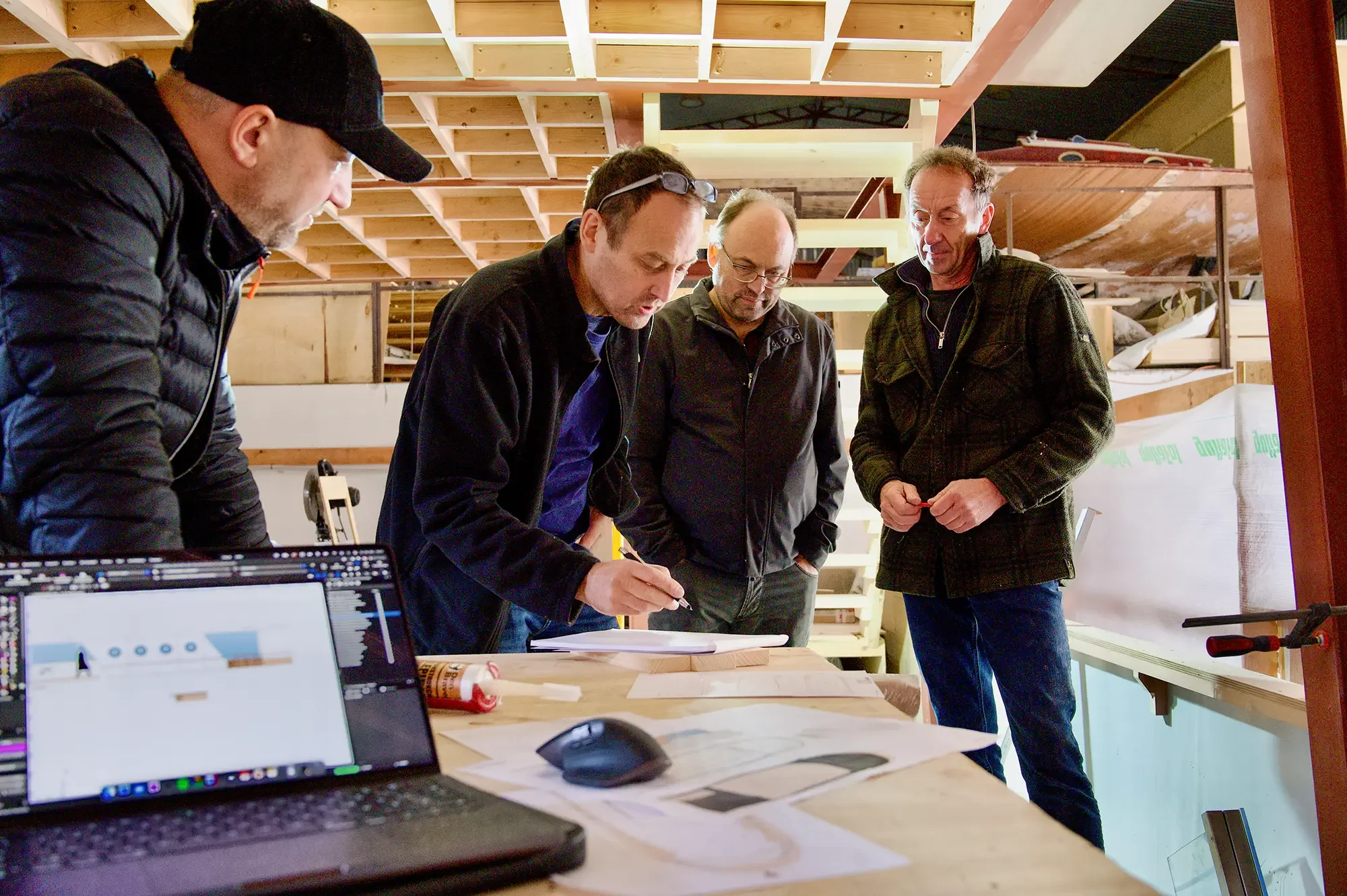 Vier Männer stehen um einen Holztisch, auf dem technische Zeichnungen und ein Computer liegen, in einem Holzwerkstatt oder -atelier, die miteinander diskutieren.