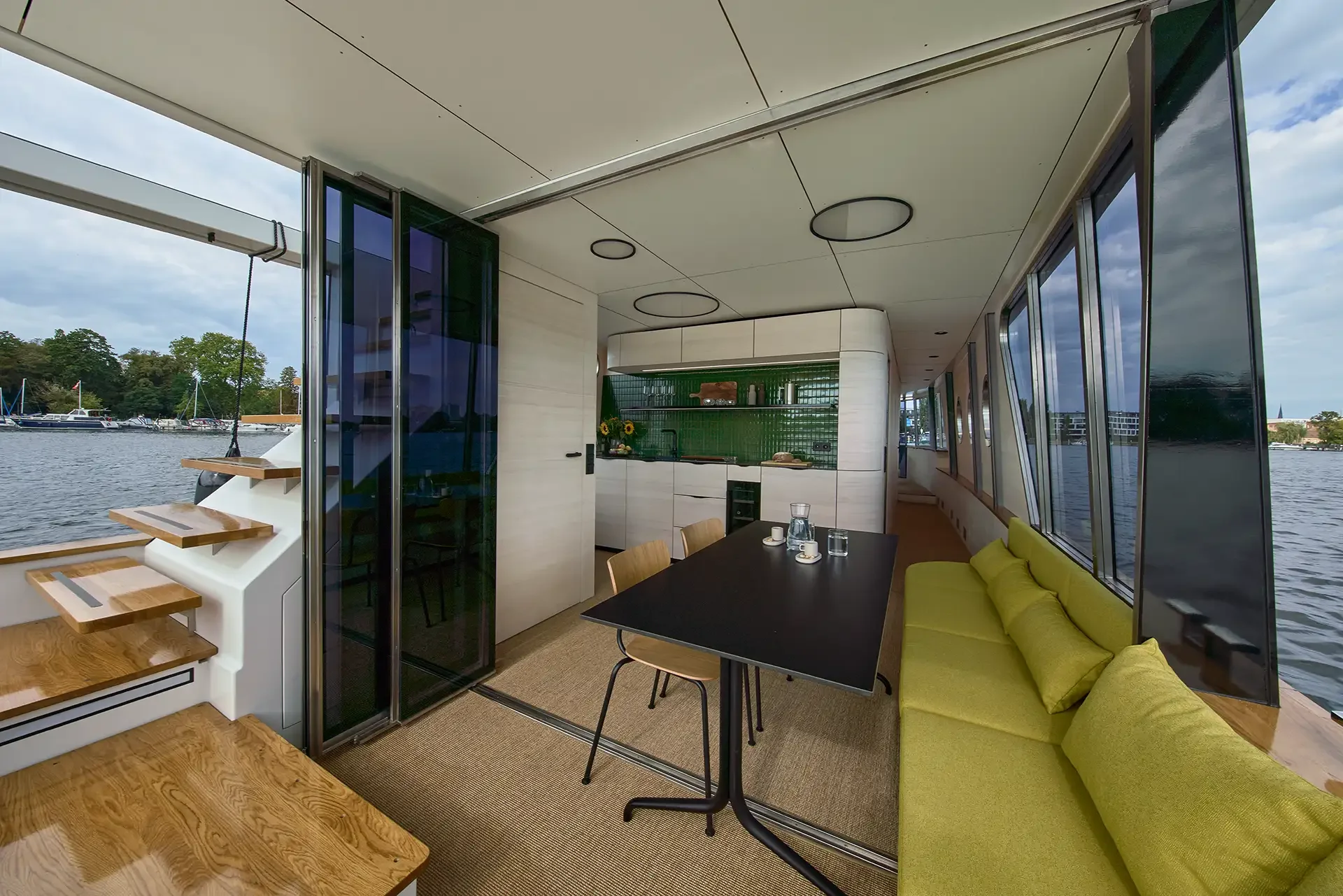 Innenansicht eines modernen Hausboots mit Blick auf den Fluss, grünem Küchenrückwand, Esstisch, grüner Sitzbank, Stühlen, Treppe und großzügigen Fensterfronten.