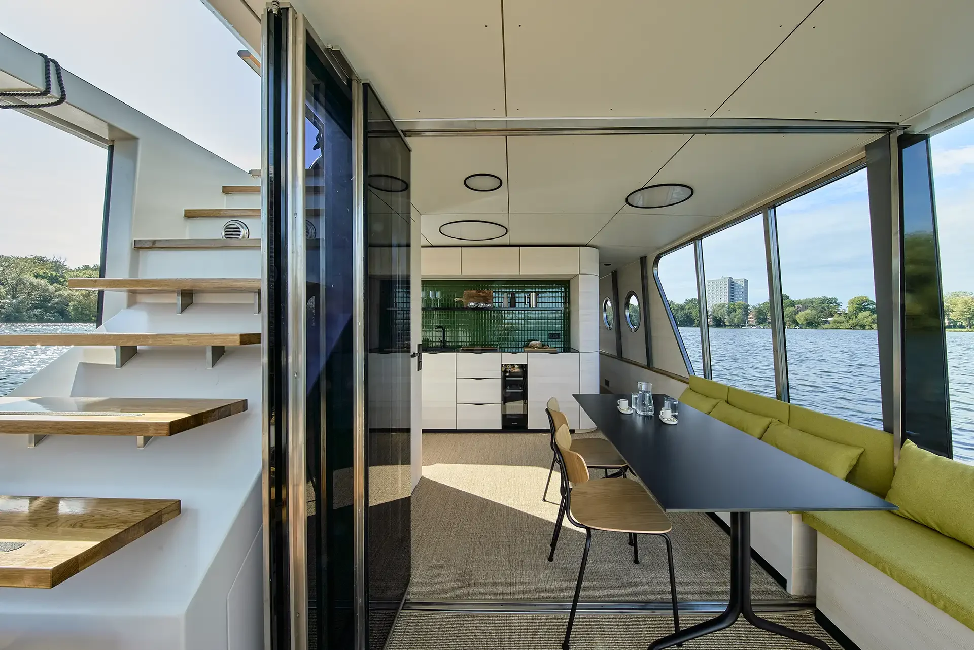 Innenansicht eines modernen Hausbootes mit Küchenbereich, Esstisch mit Stühlen, großen Fenstern und Blick auf den Fluss, moderne Einrichtung, Holzstufen, Blick nach draußen auf Wasser und Bäume.