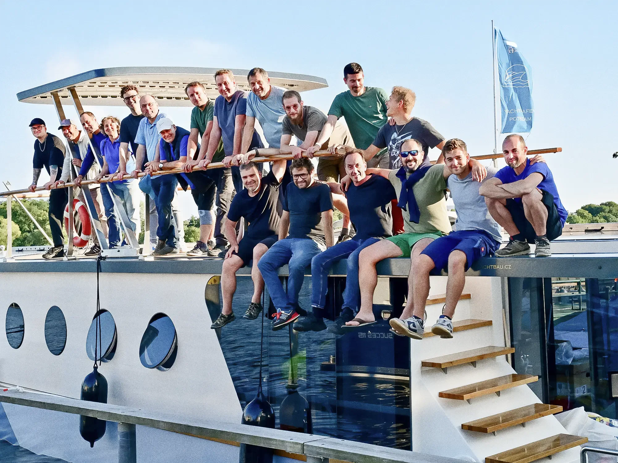 Gruppe von Menschen auf dem Deck eines Bootes, einige sitzen, andere stehen, bei Sonnenschein, einige winken, im Hintergrund Bäume und blauer Himmel.