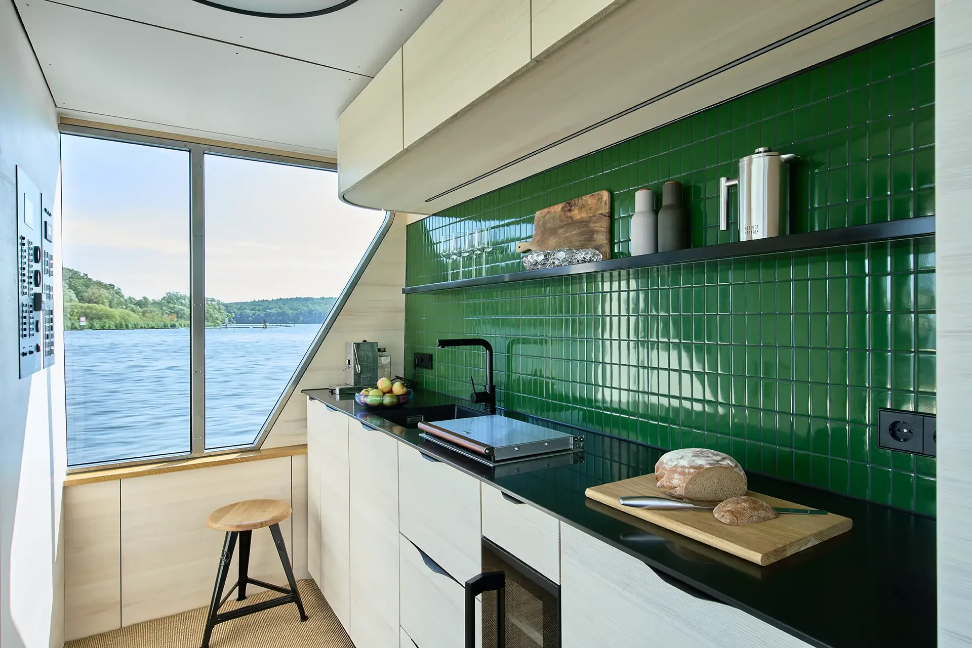 Moderne Küche mit grüner Fliesenspiegel, Fenster mit Blick auf einen Fluss und grüne Landschaft, Küchenarbeitsplatte mit Obst, Brotaufschnitt und Geschirr.