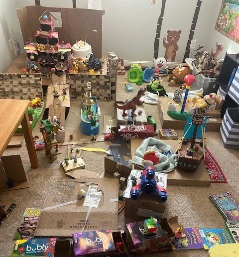 Child's playroom with various toys, including a pink and black dollhouse, stuffed animals, action figures, and a train set, with some toys scattered across the floor and on furniture.