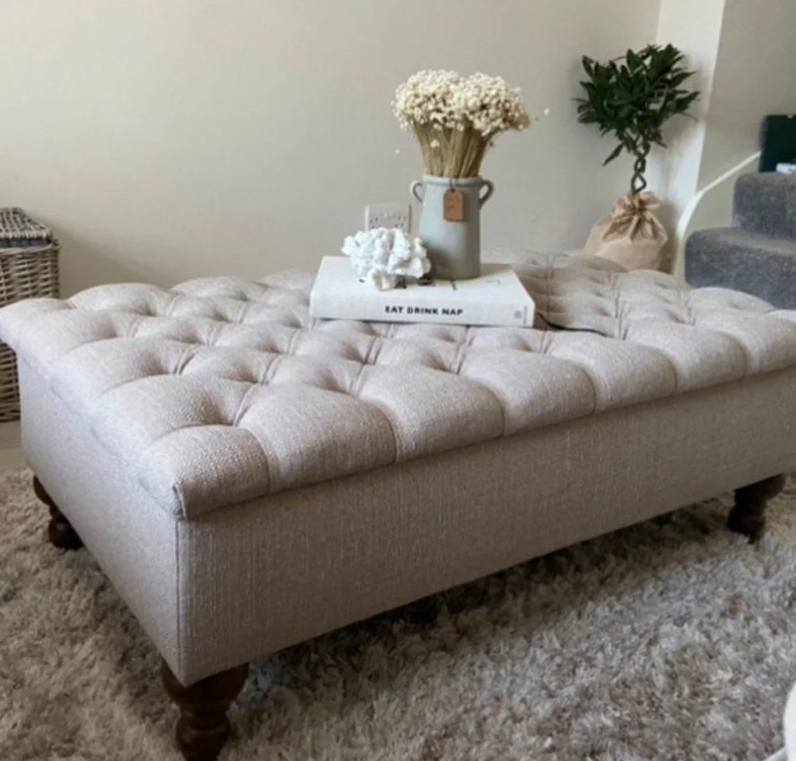 A beige tufted upholstered bench with dark wooden legs, placed on a cream shaggy rug. On top of the bench is a white tray with the words "EAT DRINK NAP" written on it, holding a clear vase with white dried flowers, a white coral sculpture, and a small white object. In the background, there's a wicker basket, a green potted plant wrapped in beige fabric, and part of a staircase with gray carpet.