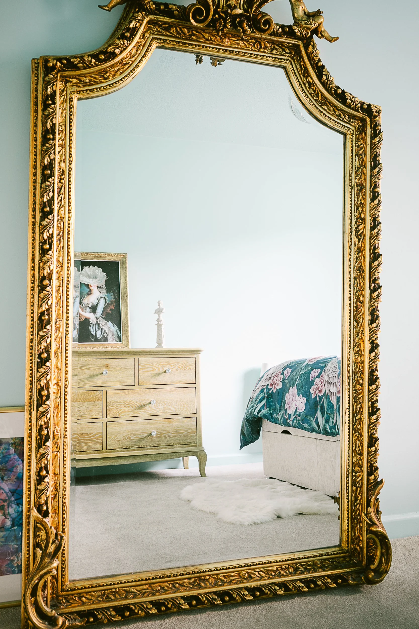 A large ornate gold-framed mirror reflecting a bedroom with a wooden dresser, a framed portrait, a small statuette, a bed with floral bedding, a white fluffy rug, and light-colored walls.