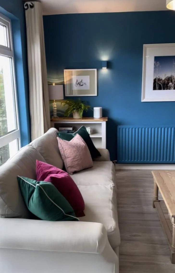 Living room with beige sofa, pink, green, and black throw pillows, blue wall, white framed artwork, small white bookshelf with a potted fern, table lamp, and window with beige curtains.