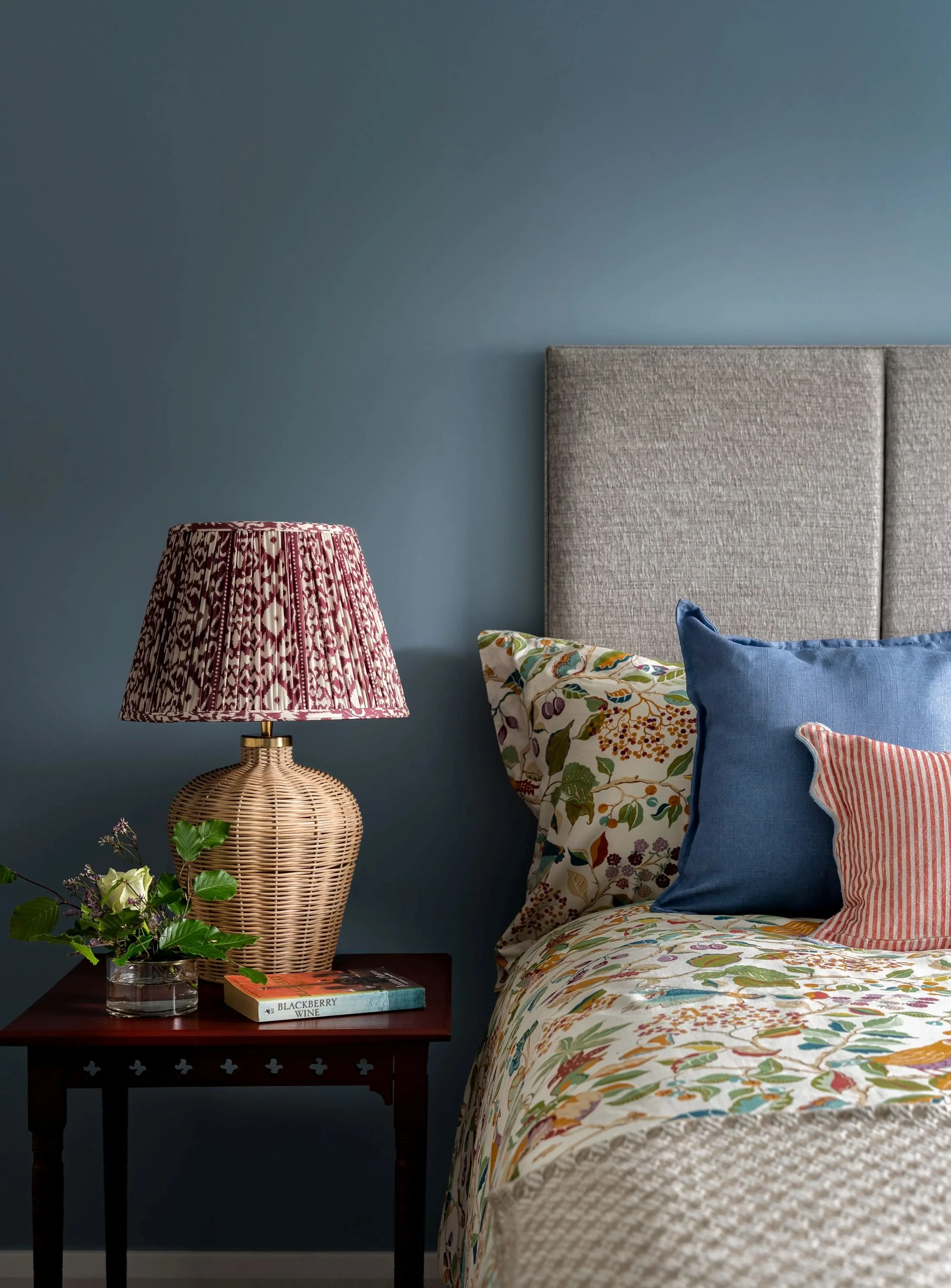 Country-inspired bedroom with smokey blue walls, wicker lamp base and colourful pattern.