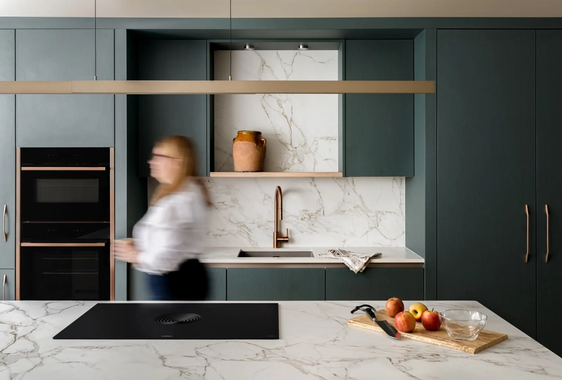 Modern kitchen with blue cabinetry, white marble countertops, and a marble backsplash, featuring a copper faucet and a gold-colored hanging light