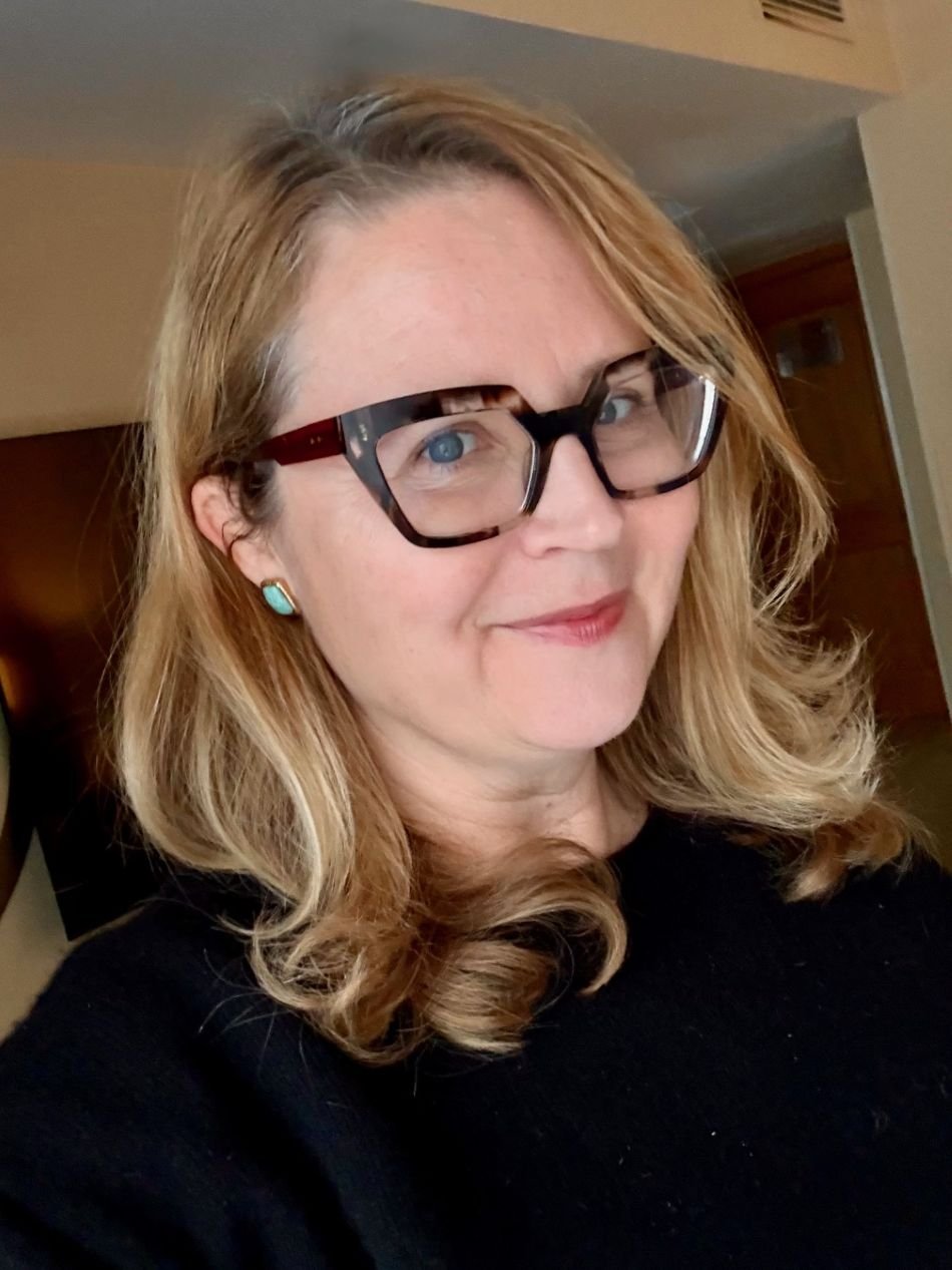 A woman with blonde hair, glasses, and turquoise earrings, wearing a black top, smiling softly at the camera indoors.