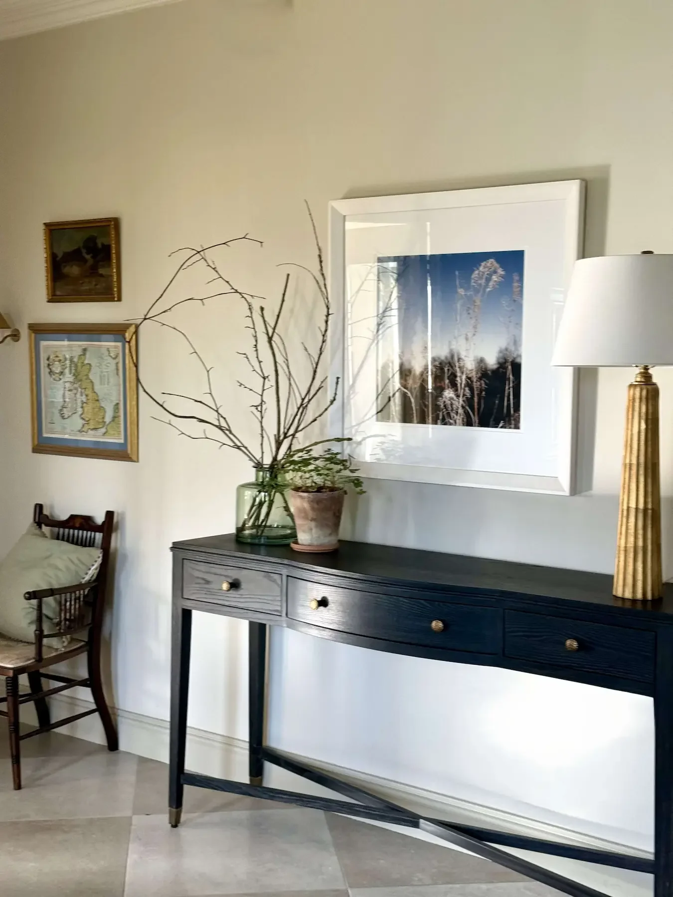 Living room with a black console table holding a large glass vase with branches and a small potted plant, a framed photograph on the wall, and a lamp with a yellow textured base.