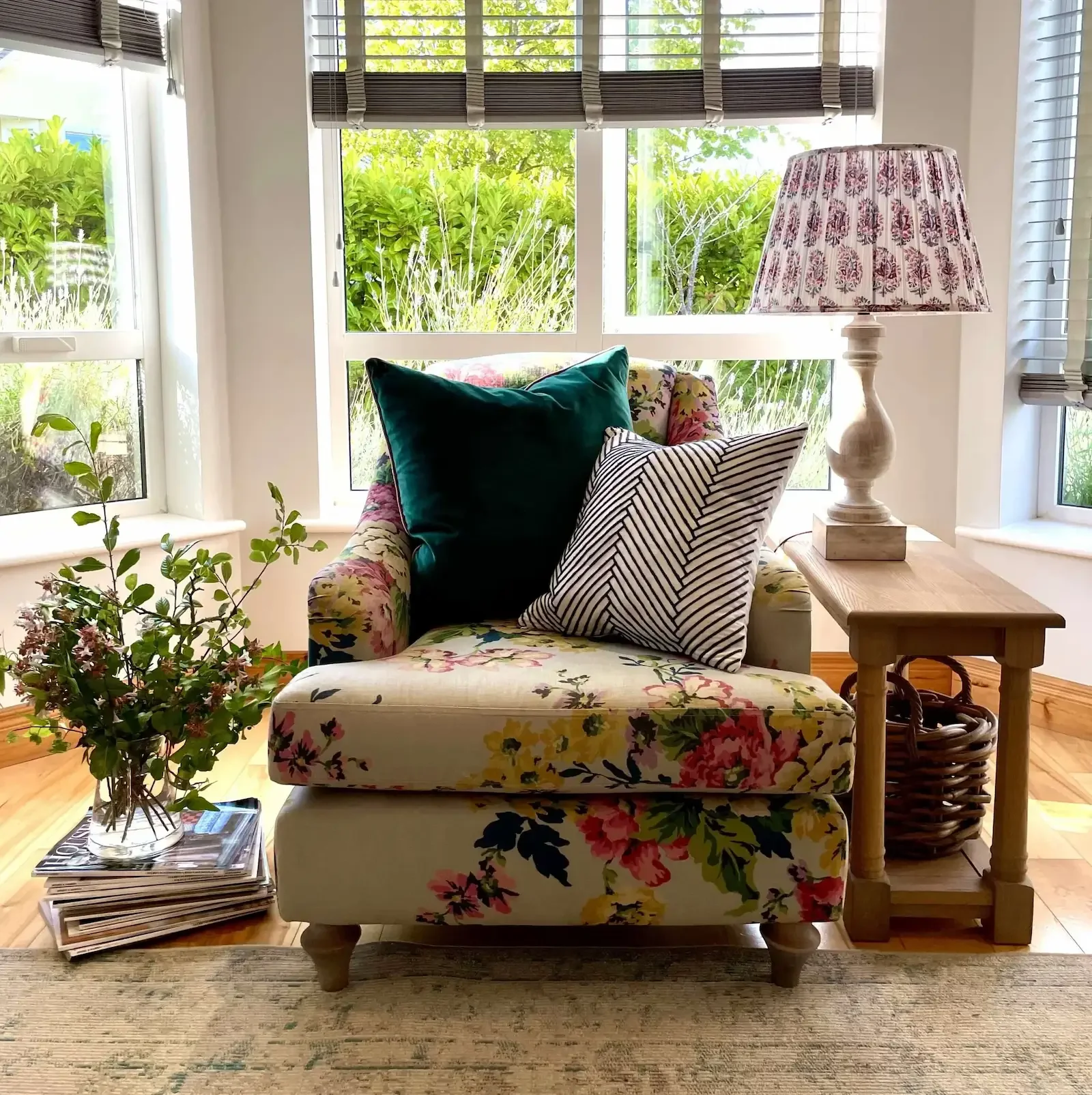 A cozy living room corner featuring a floral-patterned armchair with two pillows, a side table with a decorative lamp, and a potted plant on a small stack of magazines, all set against a backdrop of large windows with green foliage outside.