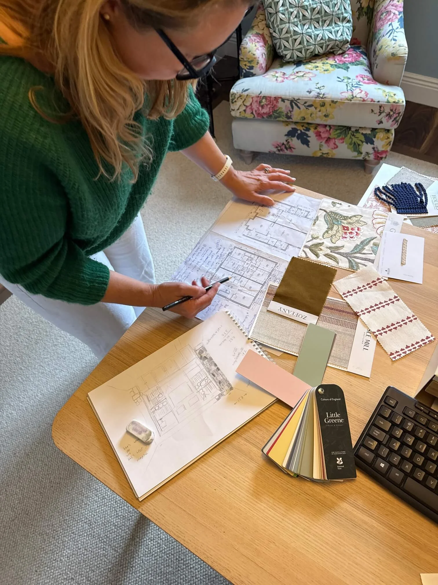 A woman in glasses and a green sweater is looking at architectural blueprints and design samples on a wooden table. There are color swatches, fabric samples, sketches, and a keyboard on the table. A floral armchair and a cushion are in the background.