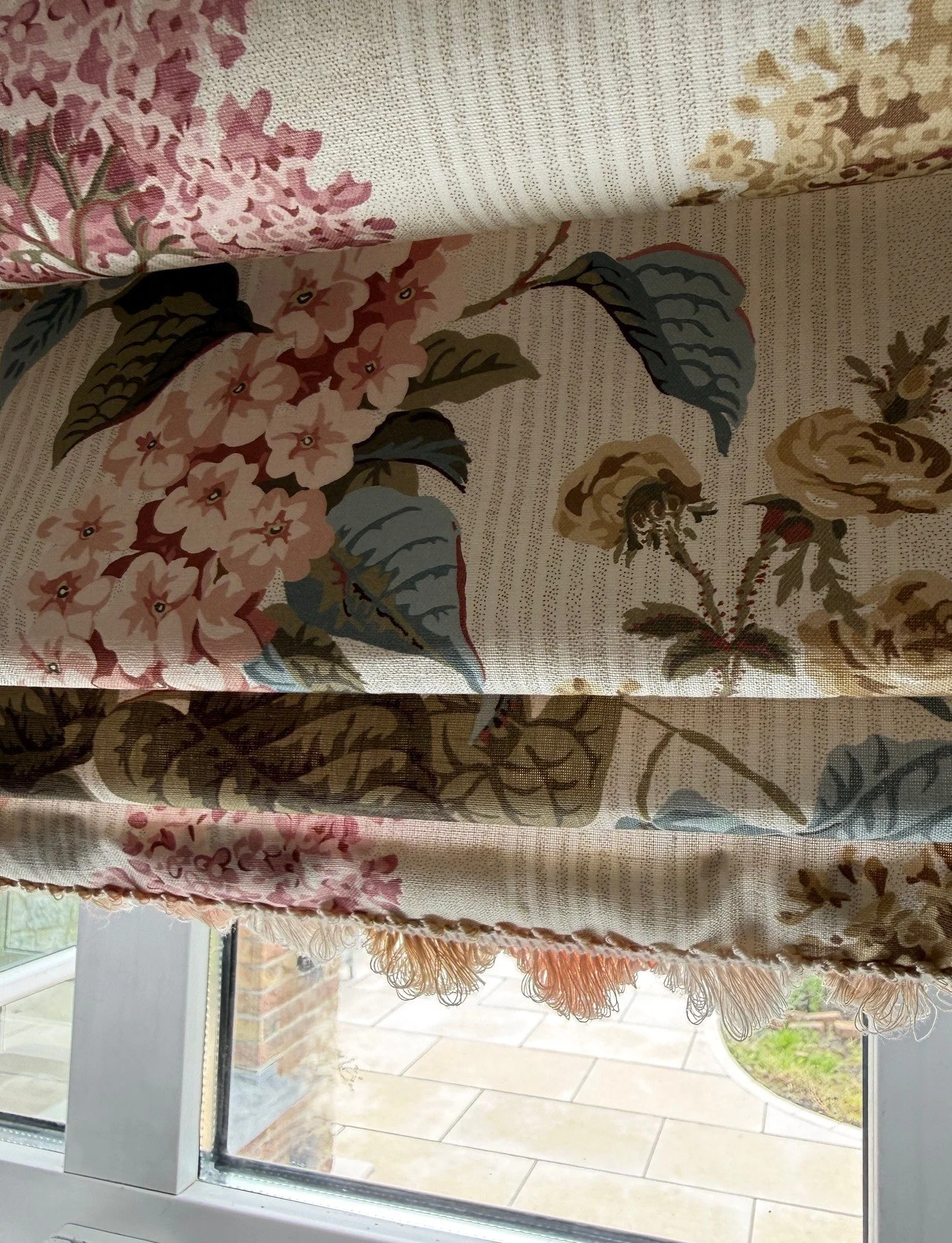 Floral Roman blind in a Sanderson fabric with pink hydrangeas, muted roses, blue-green leaves and soft fringe trim, shown lowered over a white-framed window.