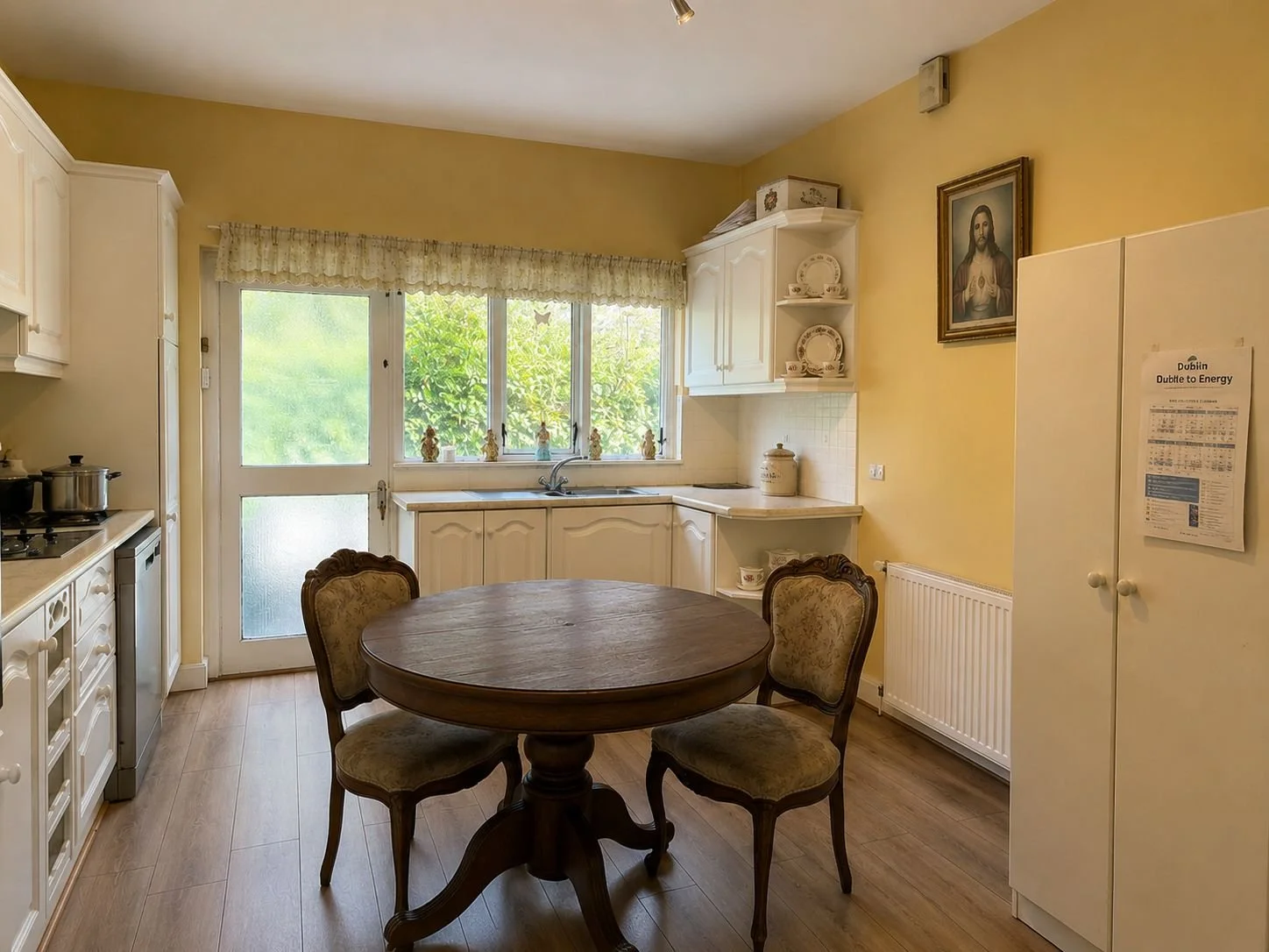 Bright kitchen with white cabinets, a wooden dining table with three upholstered chairs, yellow walls, a large window with a view of green foliage, and religious artwork.