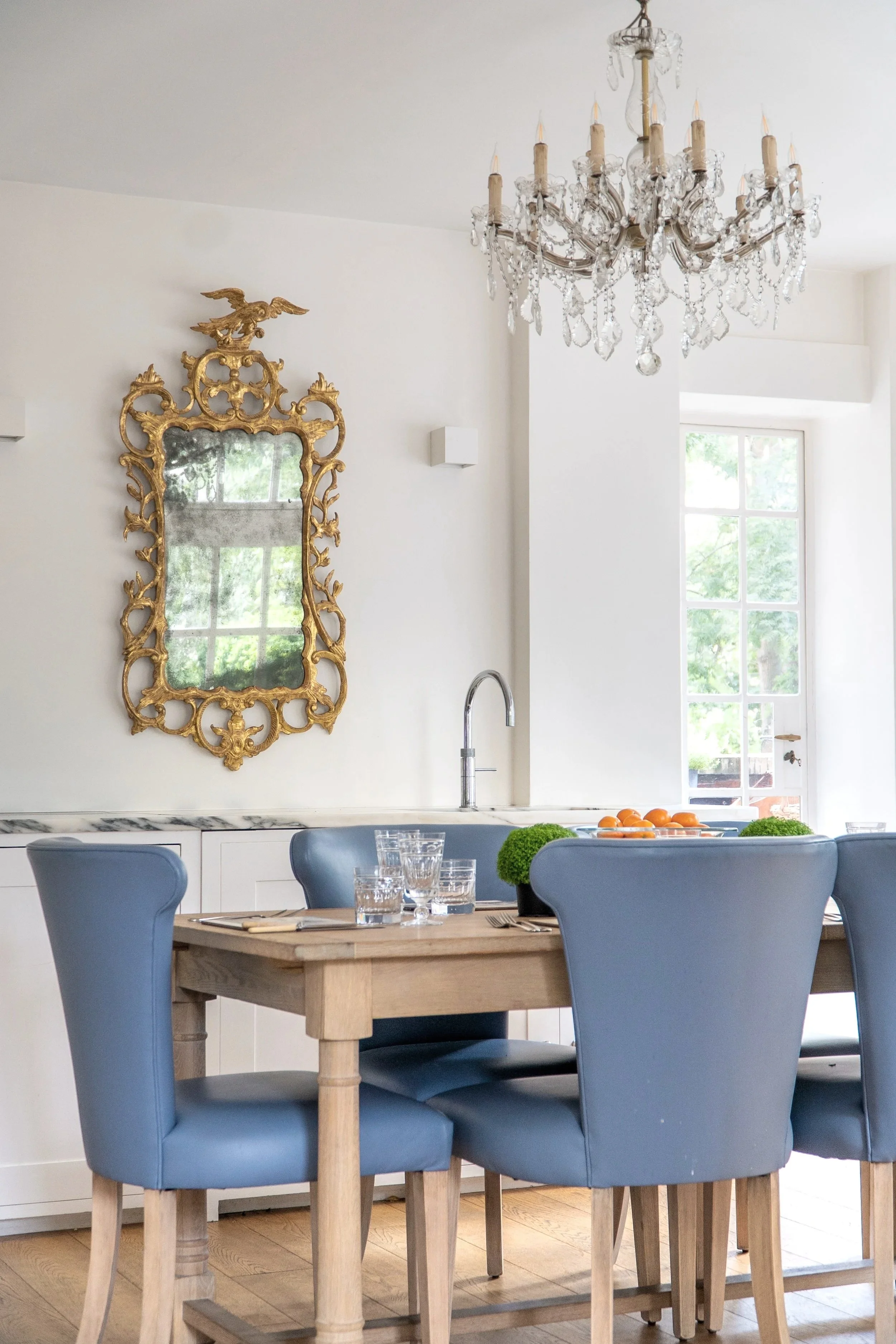 A dining room with a wooden table, blue upholstered chairs, and a crystal chandelier hanging from the ceiling. There is a large window in the background and a decorative gold mirror on the wall.