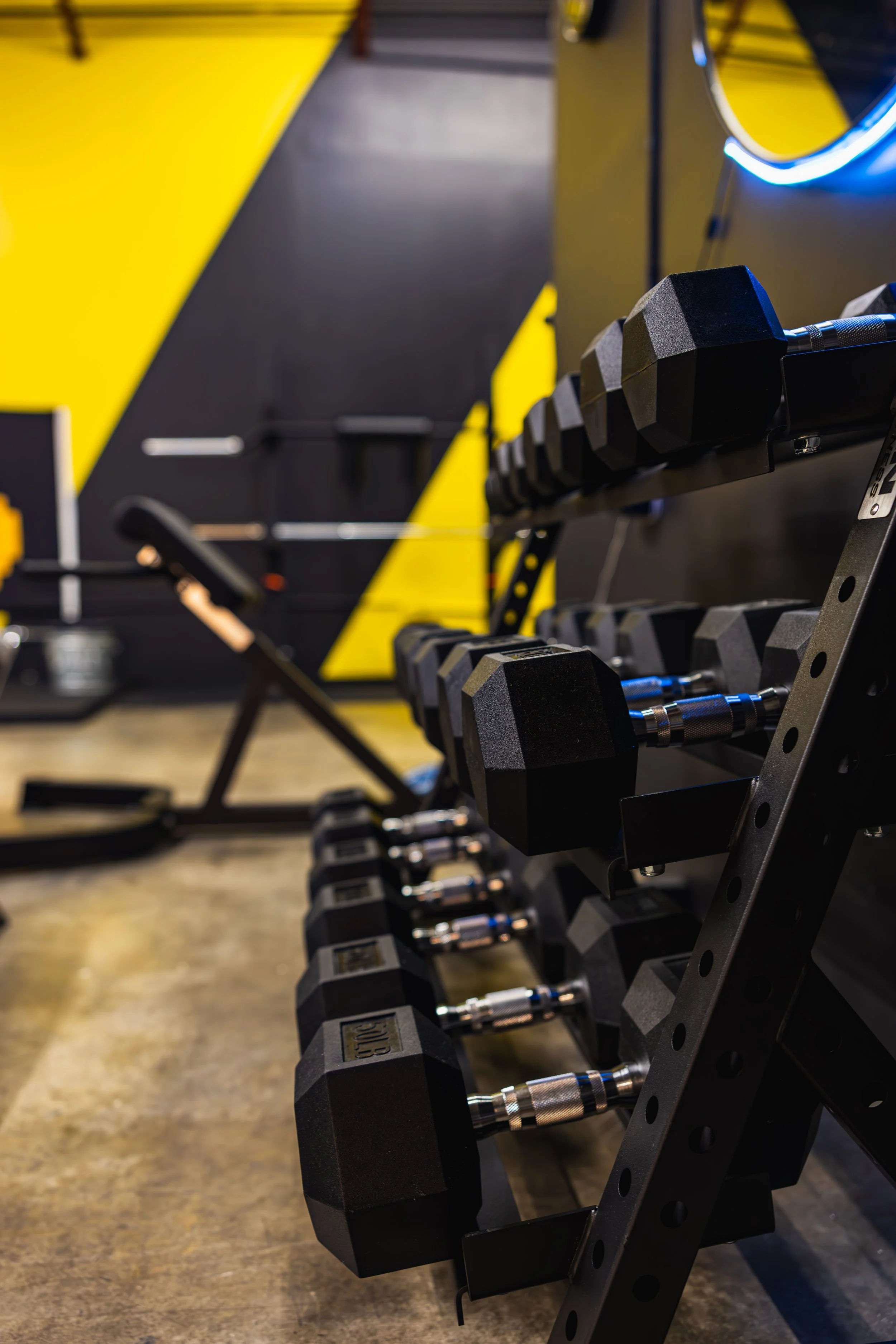 A rack of black and gray dumbbells with hexagonal heads on a metal stand inside a gym.