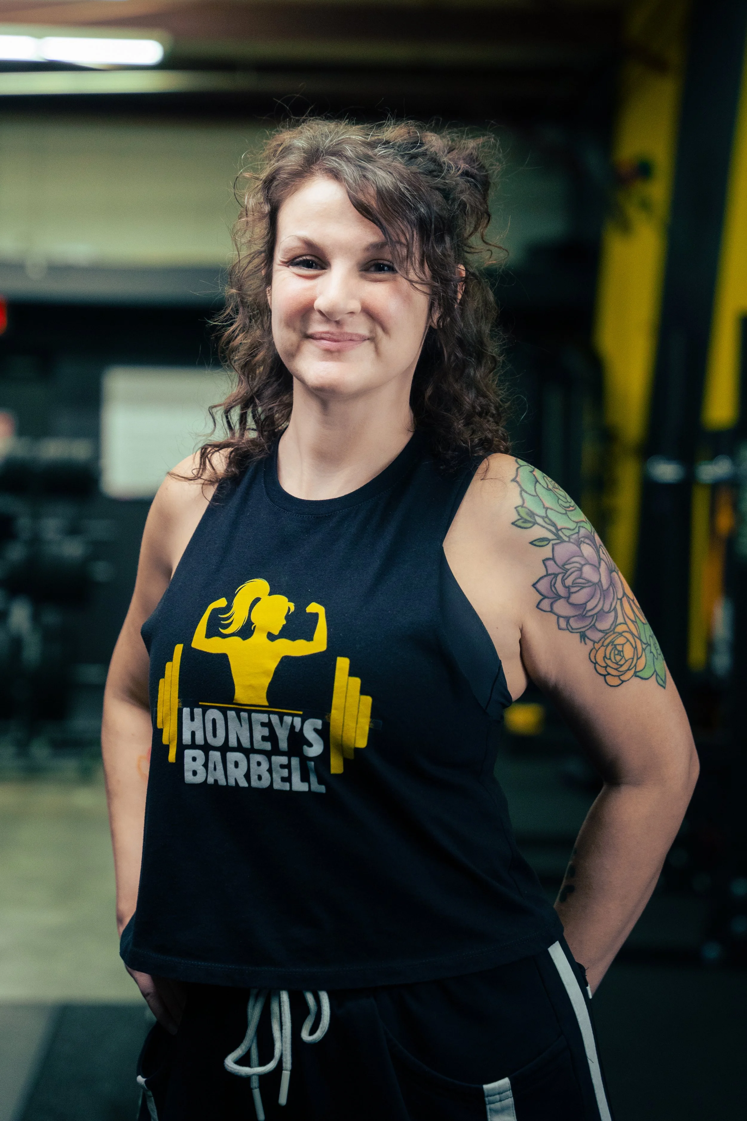 A woman with short wavy brown hair, wearing a black sleeveless athletic shirt with a yellow fitness logo, is standing against a black background. She has her arms crossed and is looking at the camera with a slight smile.