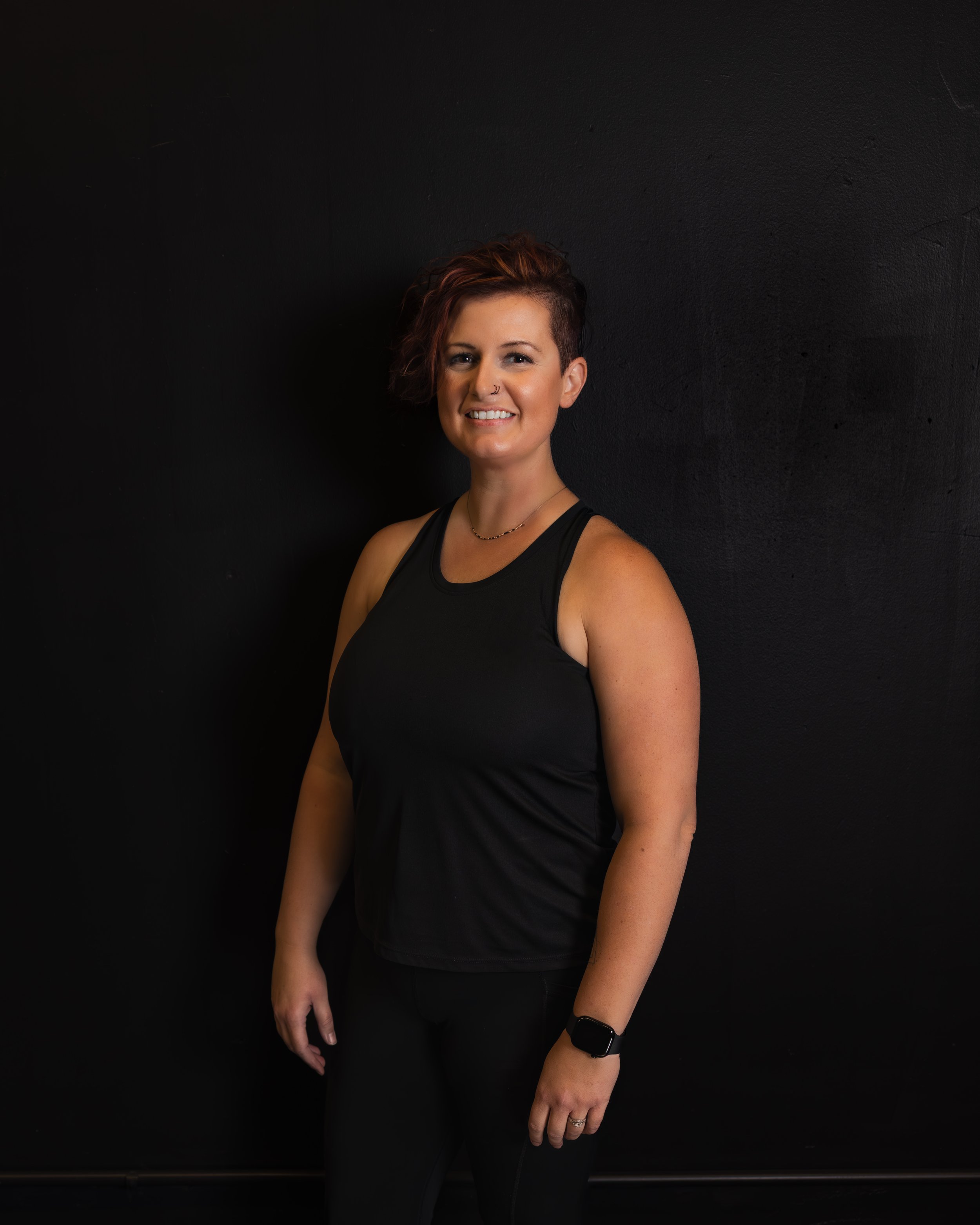 A woman with short, curly hair smiling, wearing a black tank top and black pants, standing against a dark wall.