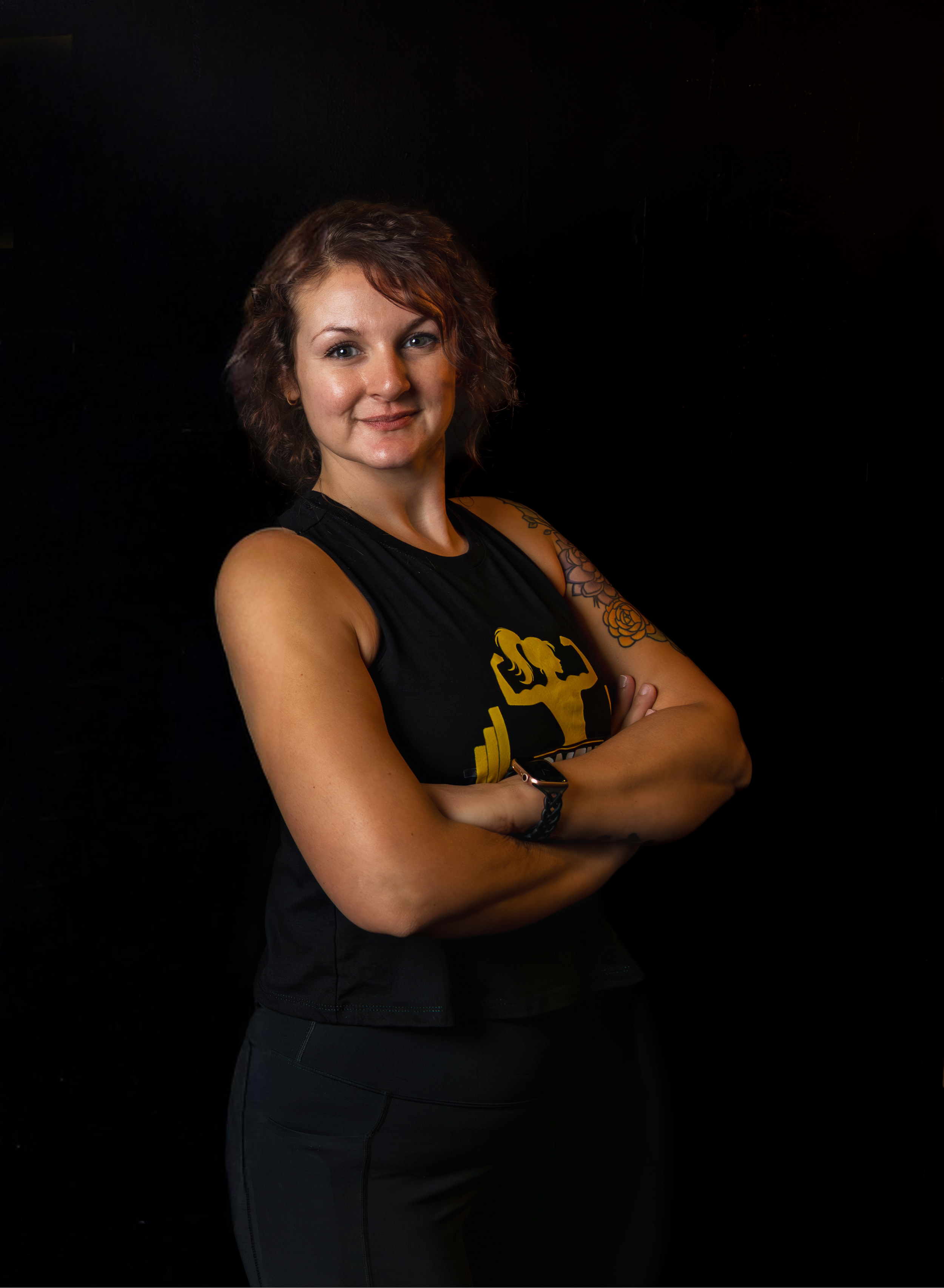 A woman with short wavy brown hair, wearing a black sleeveless athletic shirt with a yellow fitness logo, is standing against a black background. She has her arms crossed and is looking at the camera with a slight smile.